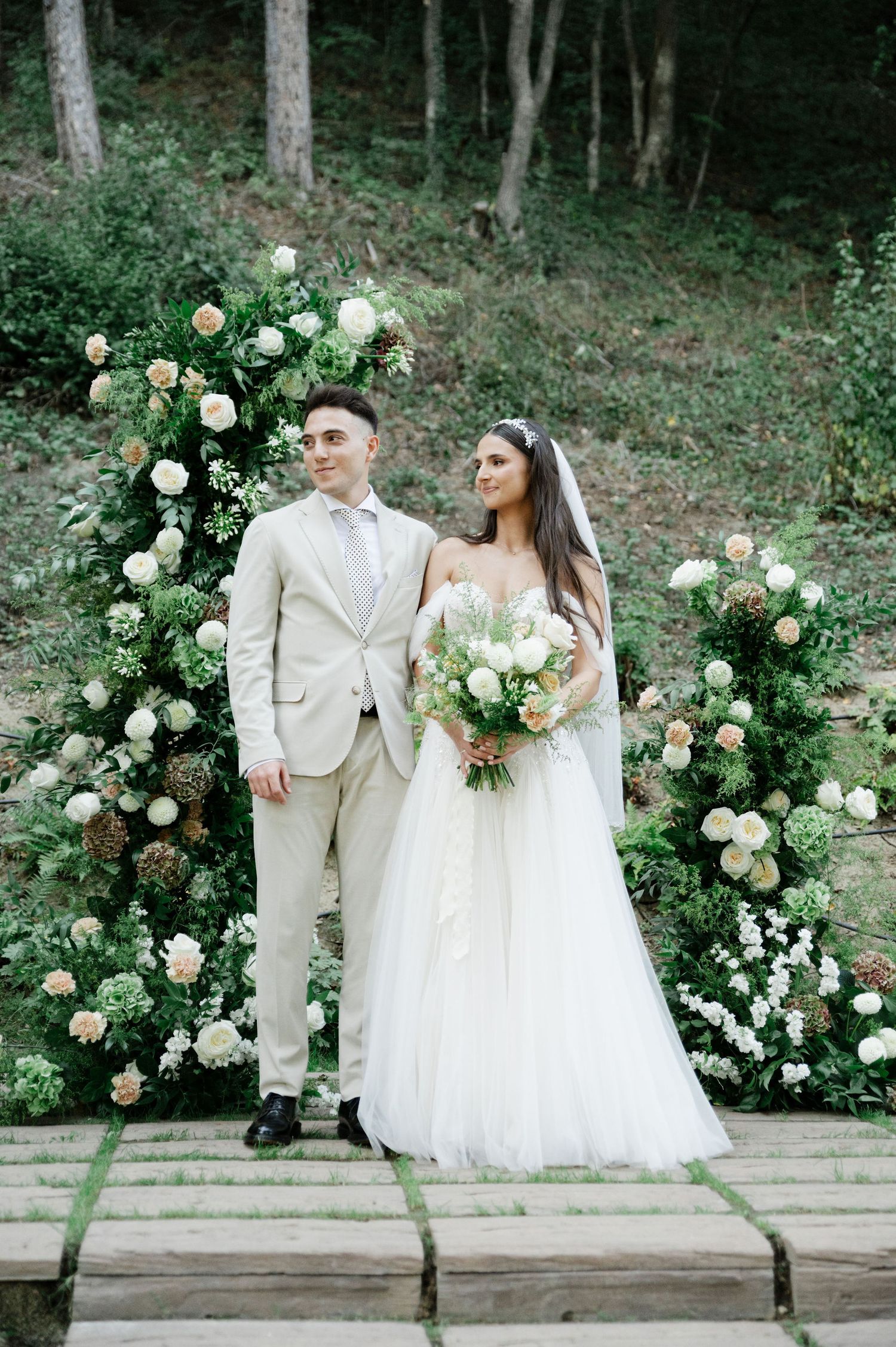 Wedding couple stands on stone platform with elegant white rose arch in forest backdrop.