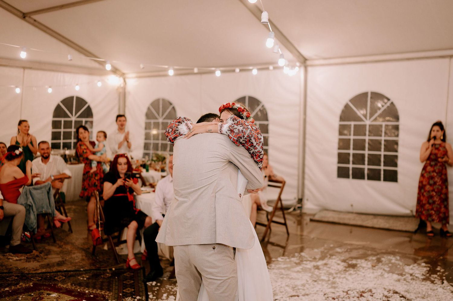 Wedding guests dance and celebrate in a tent reception venue with string lights and arched windows.