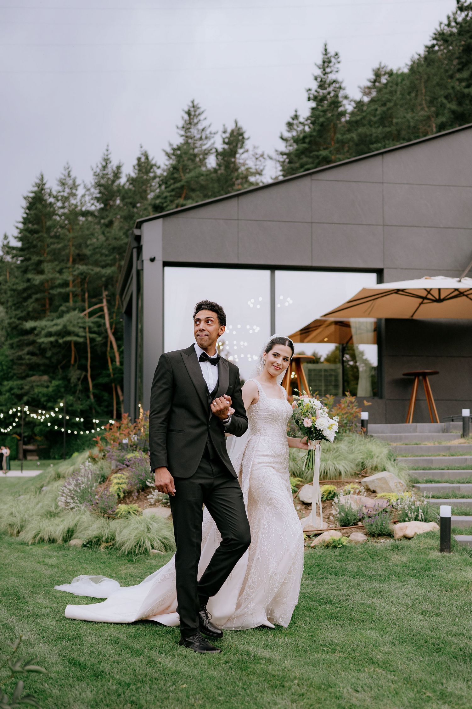 Newlyweds pose playfully in front of a modern building with string lights and landscaping.