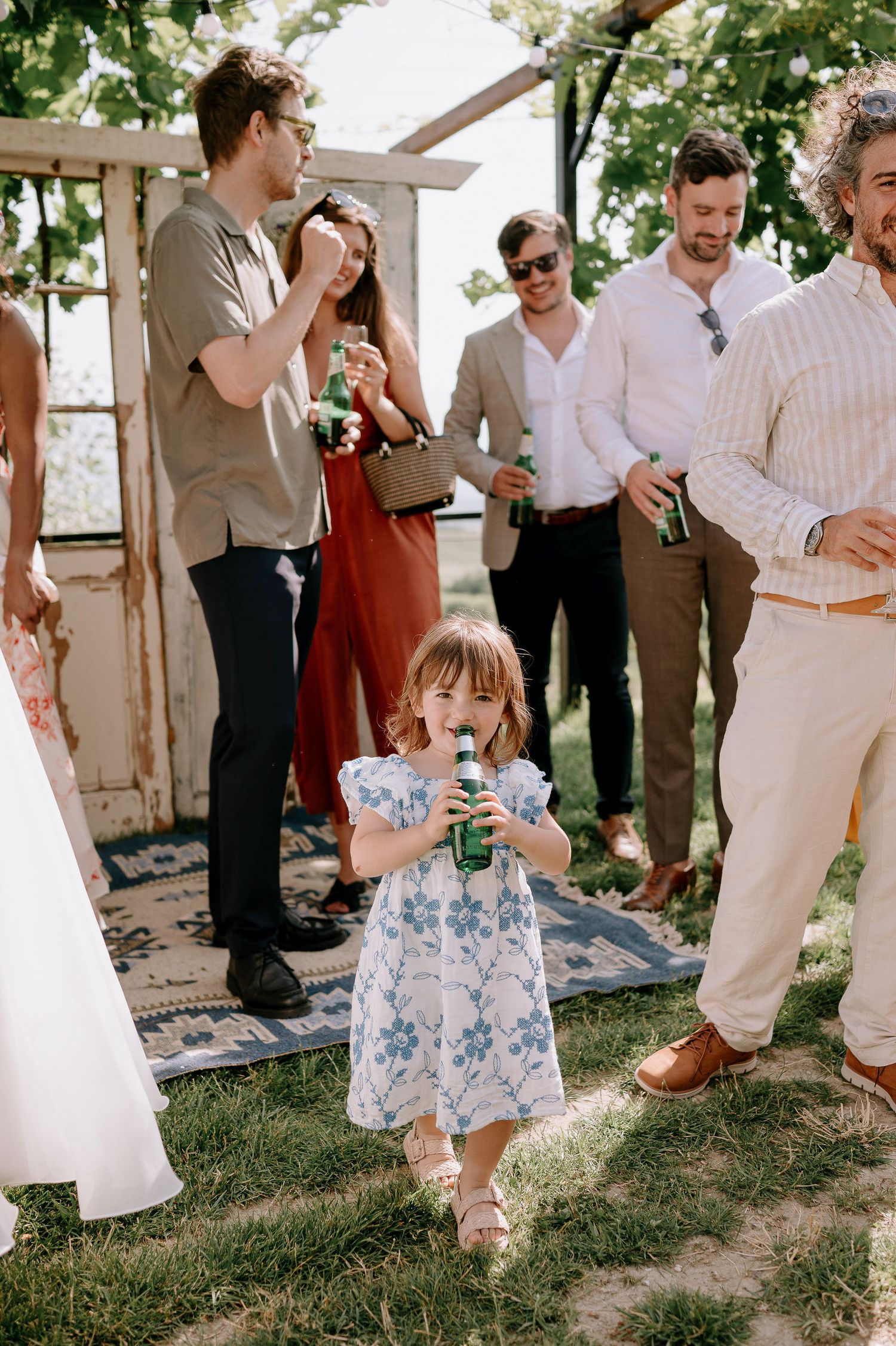 Wedding guests gather outdoors while a small child in a blue floral dress sips a beverage.