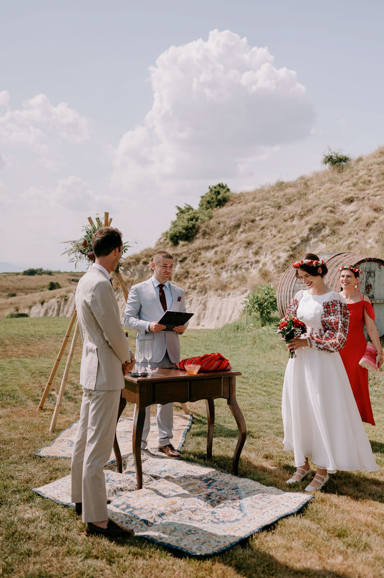 A small outdoor wedding ceremony setup with a rustic wooden table and patterned rug on a grassy hillside.