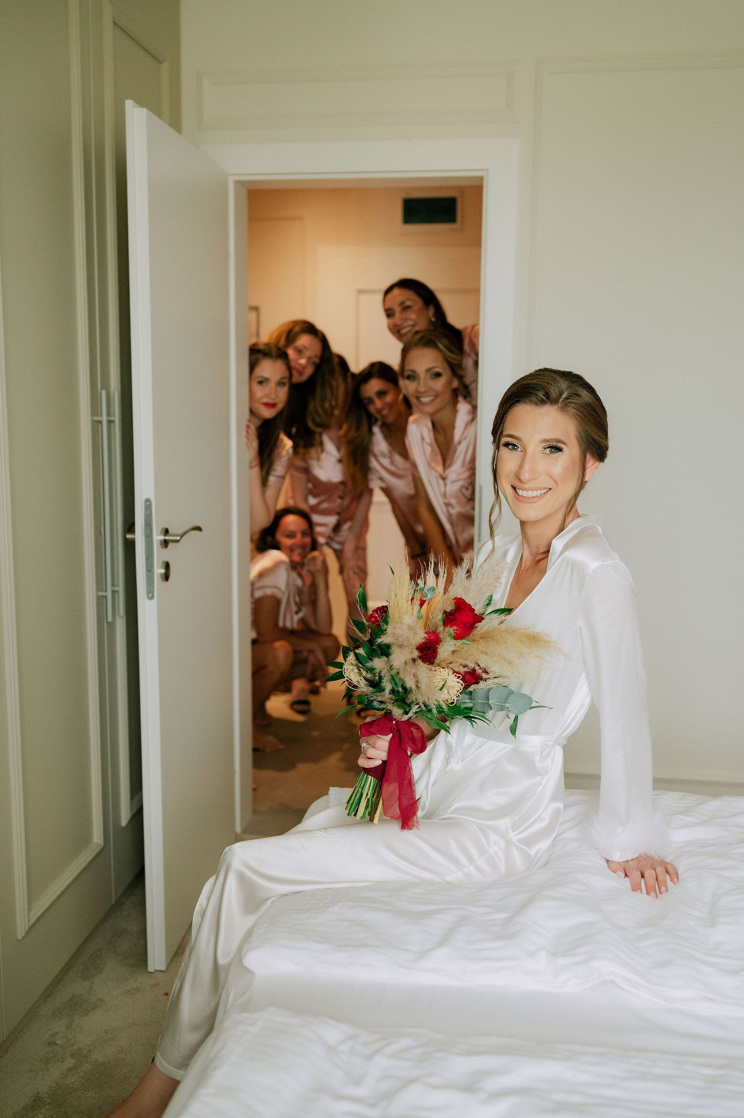 Candid wedding morning scene with bride on bed and bridesmaids reacting in doorway.