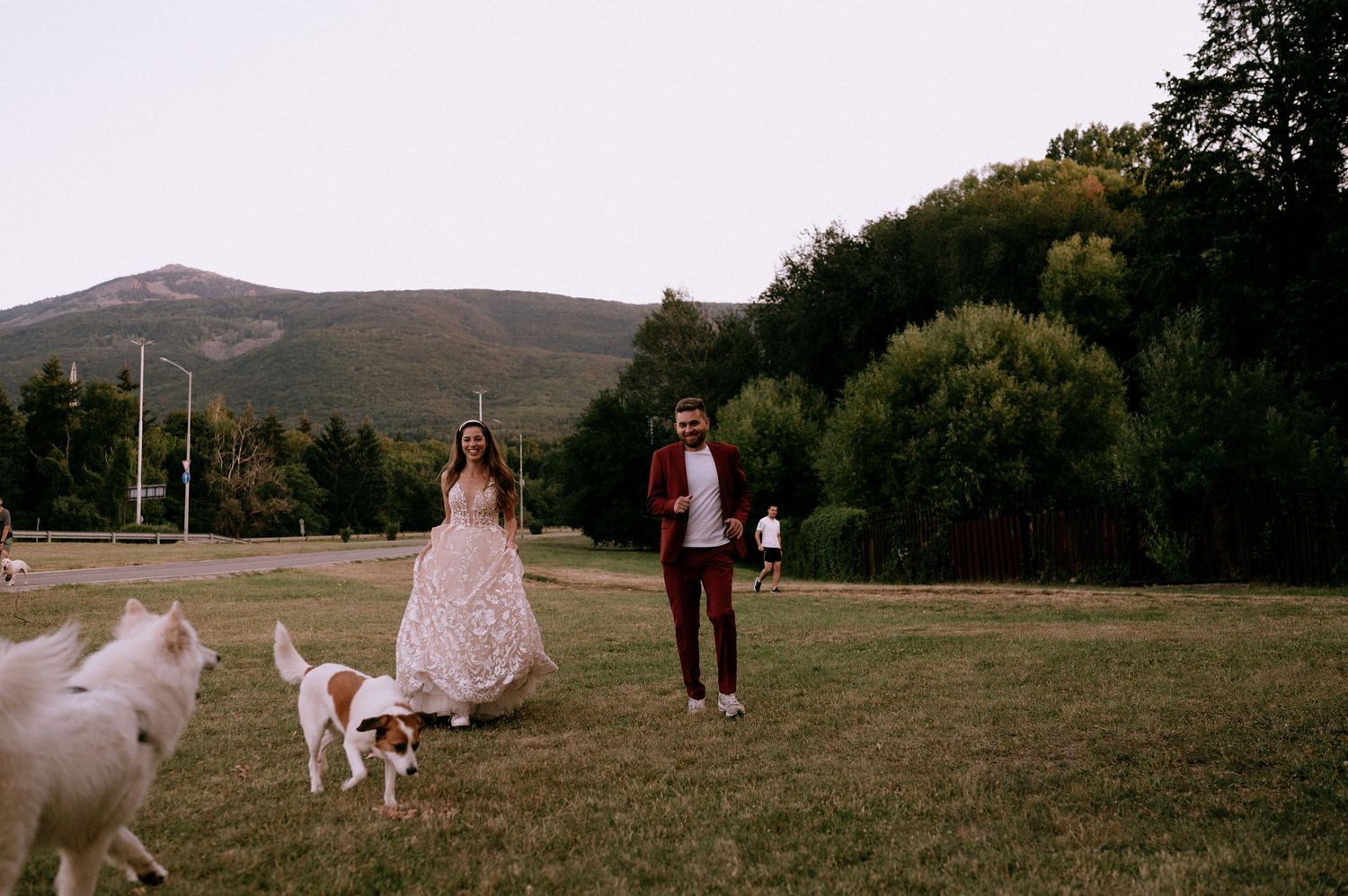 Two people and their white dog share a moment in a scenic mountain field at dusk.