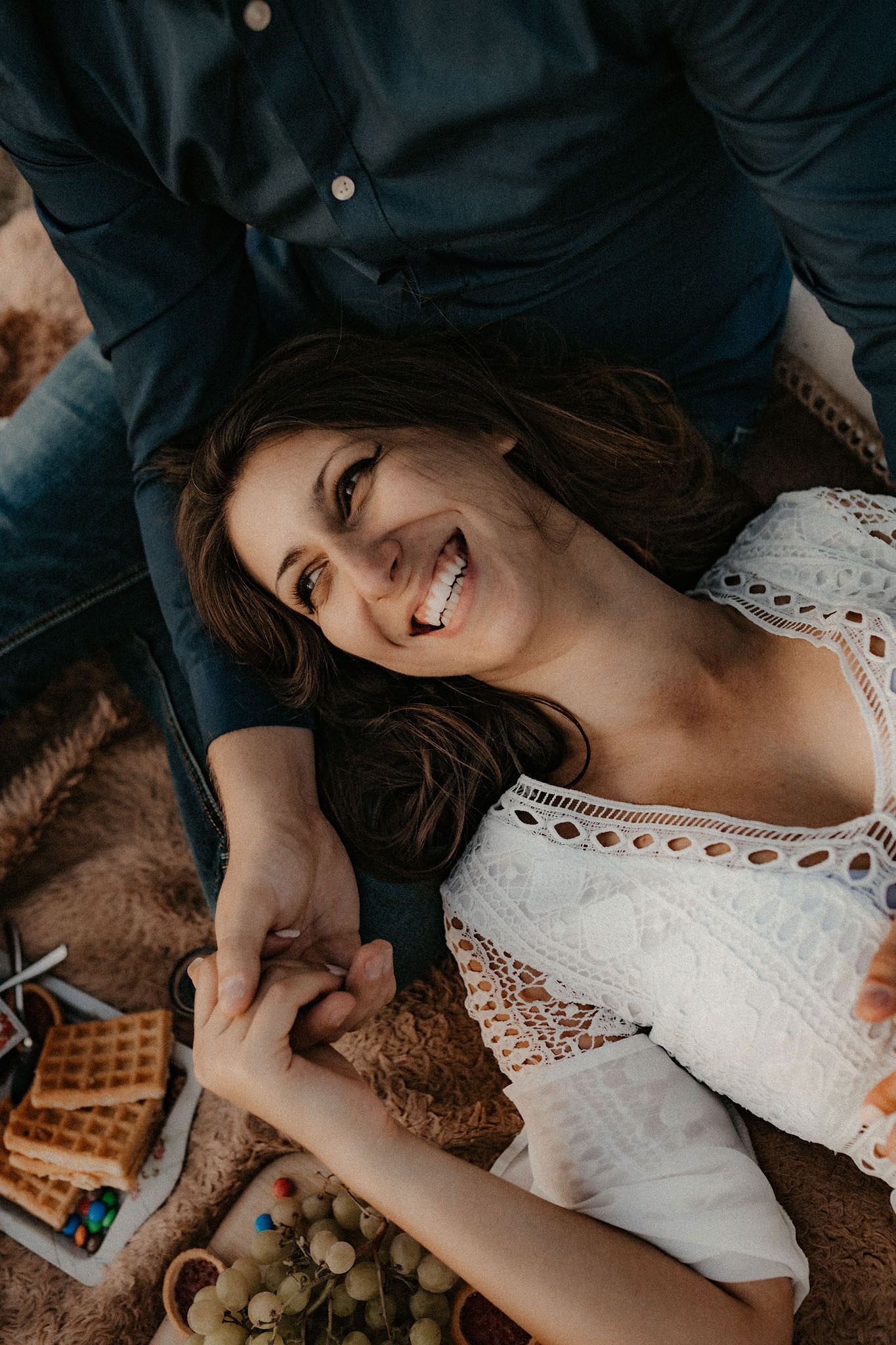 Person in white lace dress lying on blanket surrounded by picnic food and drinks.