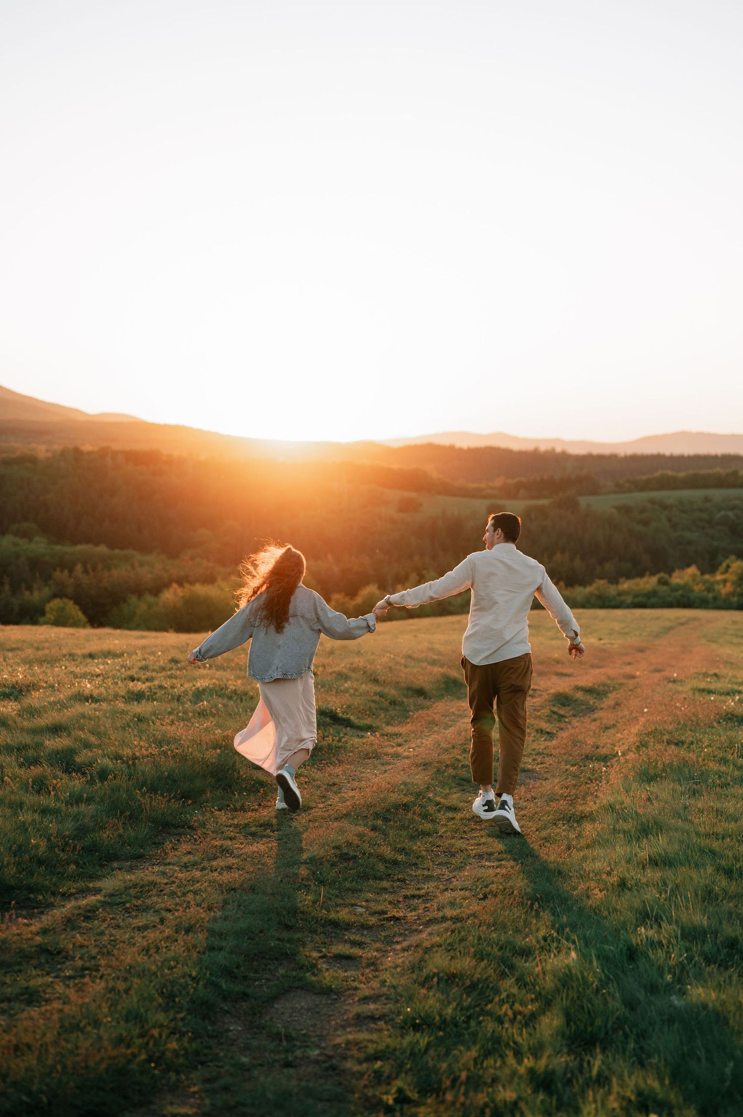 Two people holding hands and running through a grassy hillside meadow at sunset with golden light behind them.