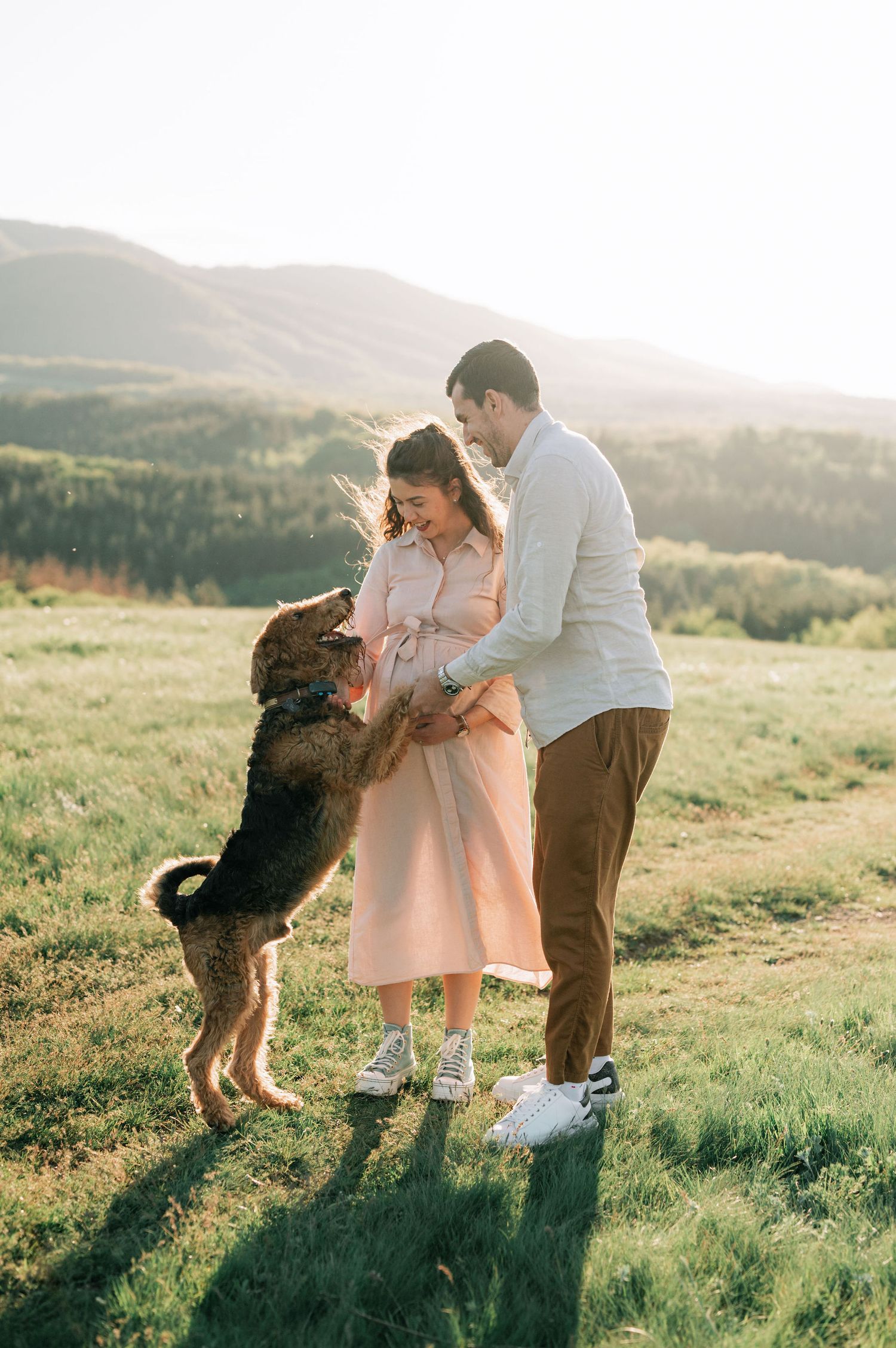 A couple shares joyful moments with their energetic dog on a grassy hilltop at sunset.
