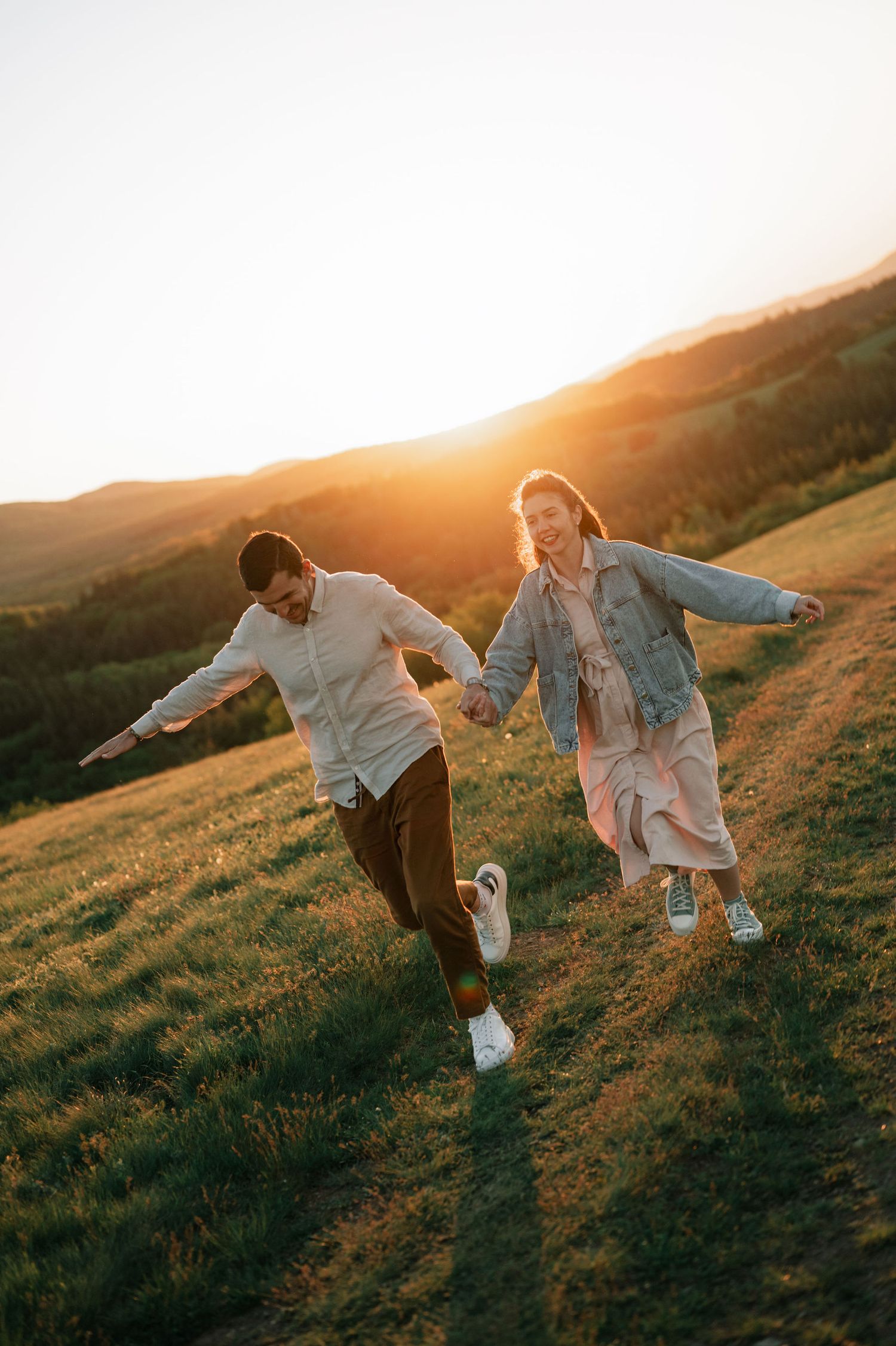 Two people in white outfits dance playfully on a grassy hillside during golden hour sunset.
