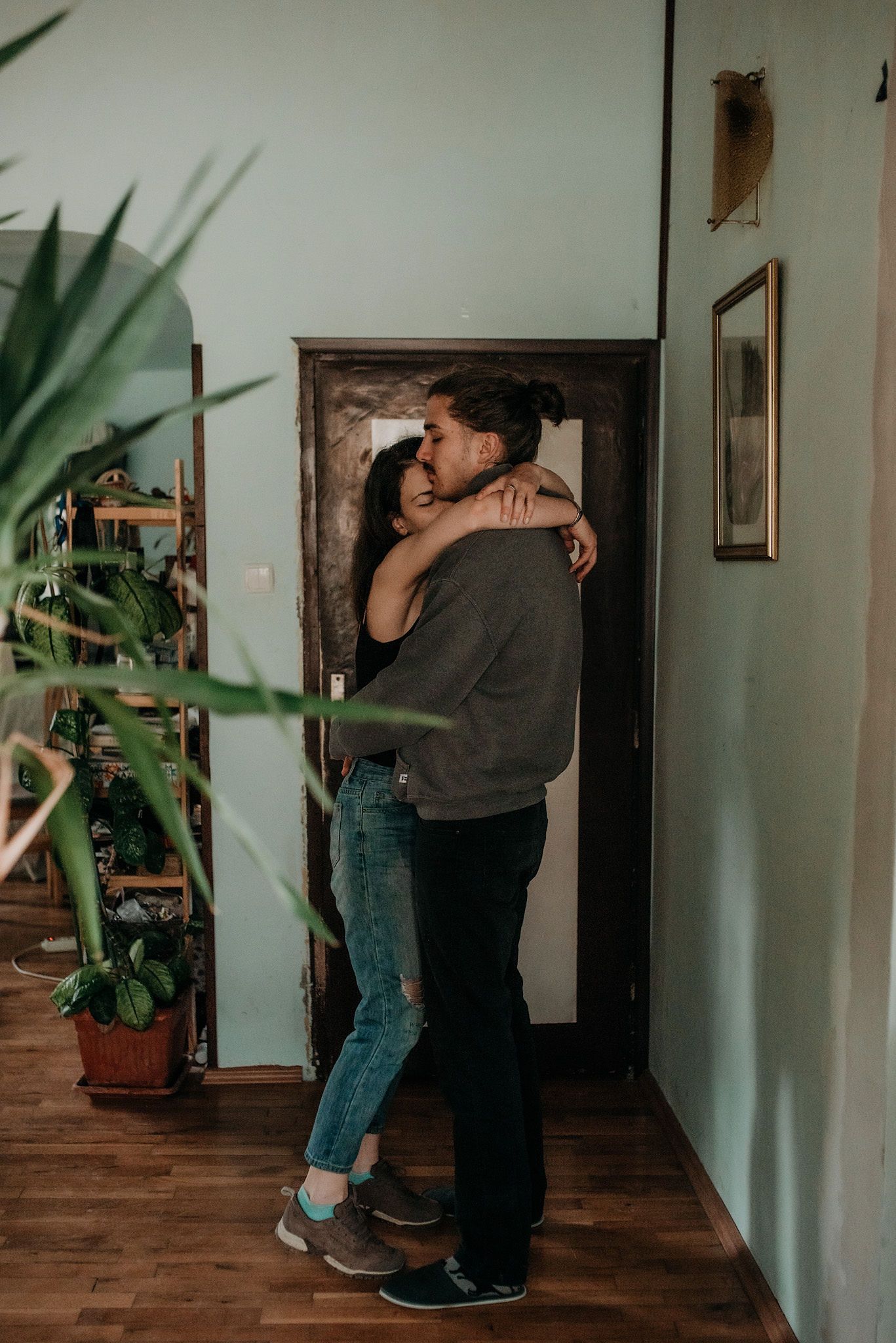 A romantic couple embraces in a cozy hallway with houseplants and wooden floors in warm lighting.