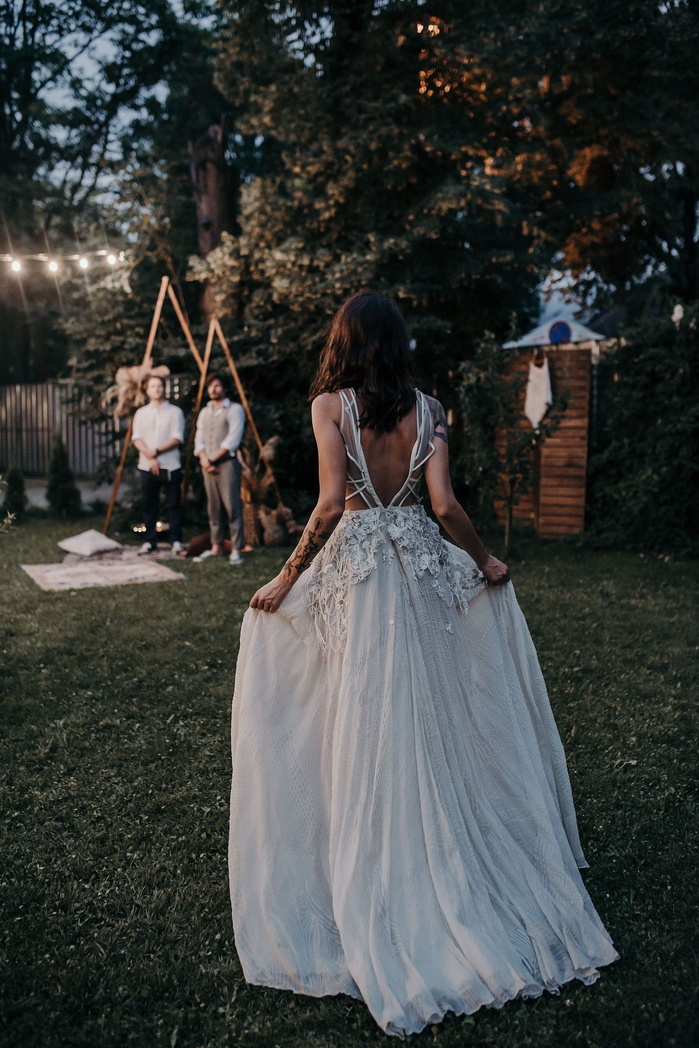Two elegant white wedding dresses flow gracefully on a lawn at dusk with string lights glowing in the background.
