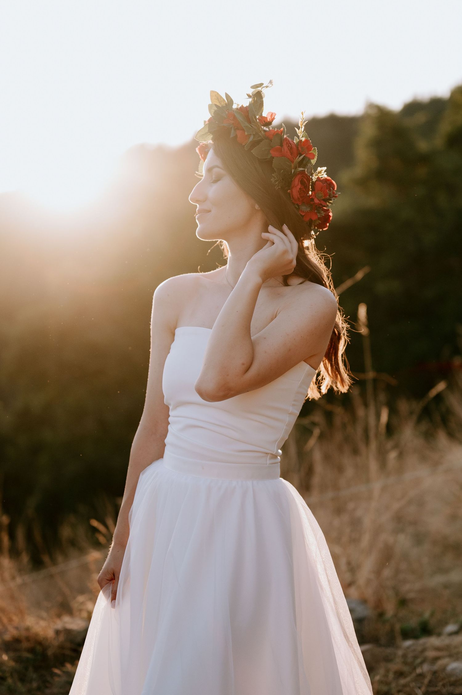 A bride in white dress and red flower crown poses in golden sunset light.