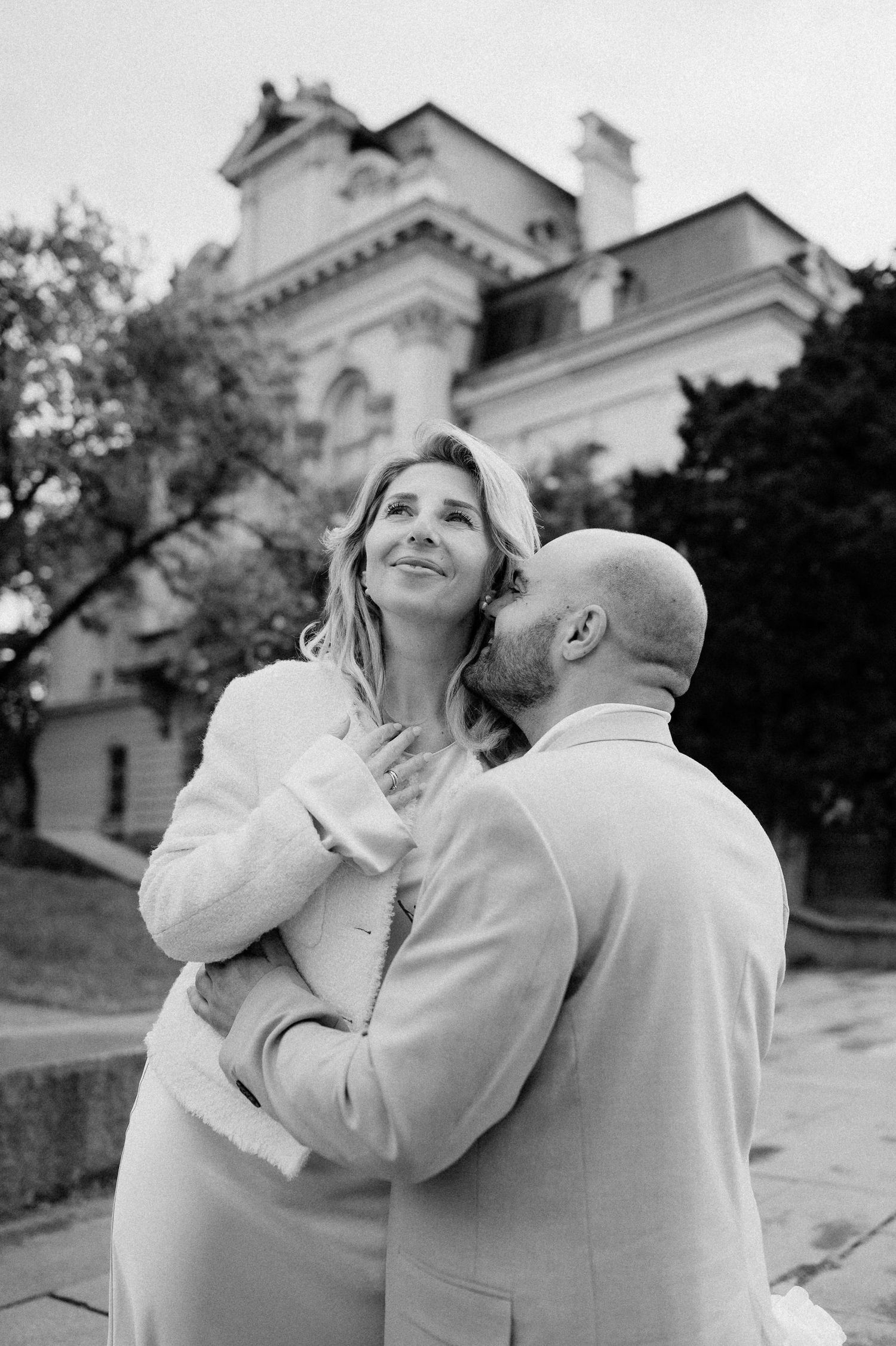 Two people in light-colored clothing embrace lovingly against the backdrop of a stately building.