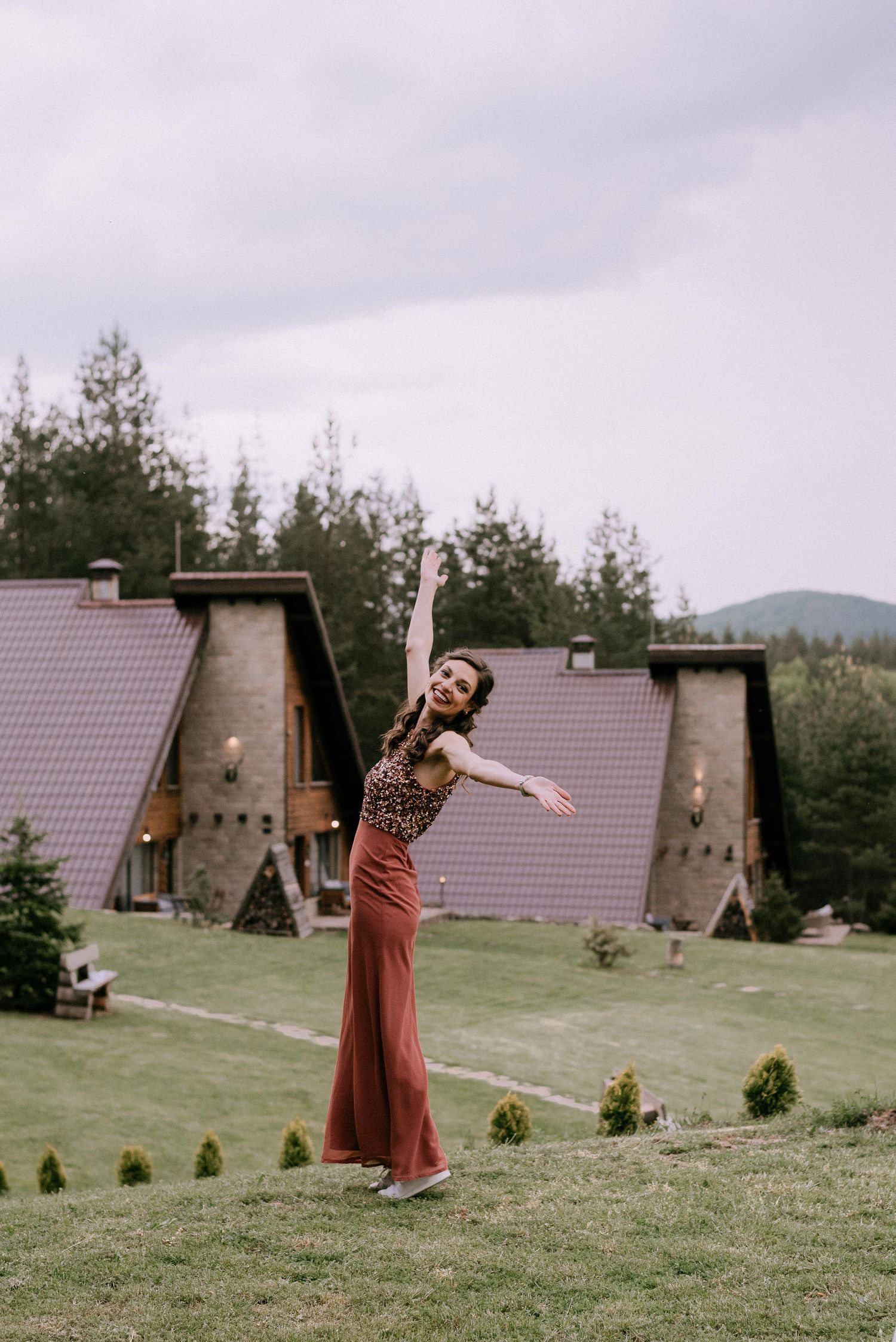 A person in a brown dress twirls joyfully on a grassy hill with A-frame cabins in the background.