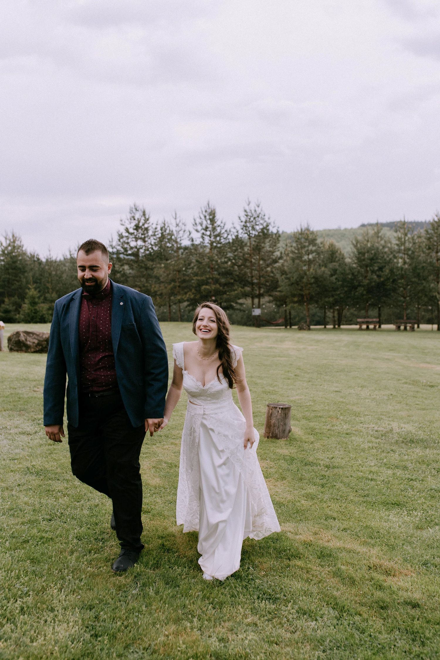 A couple laughs while walking together on grass with evergreen trees behind them.