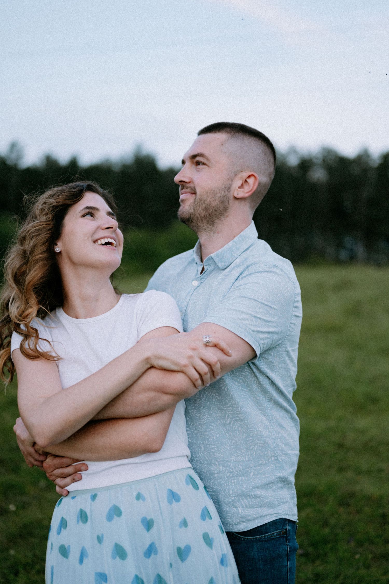 A couple shares genuine laughter while embracing in a green field at dusk.