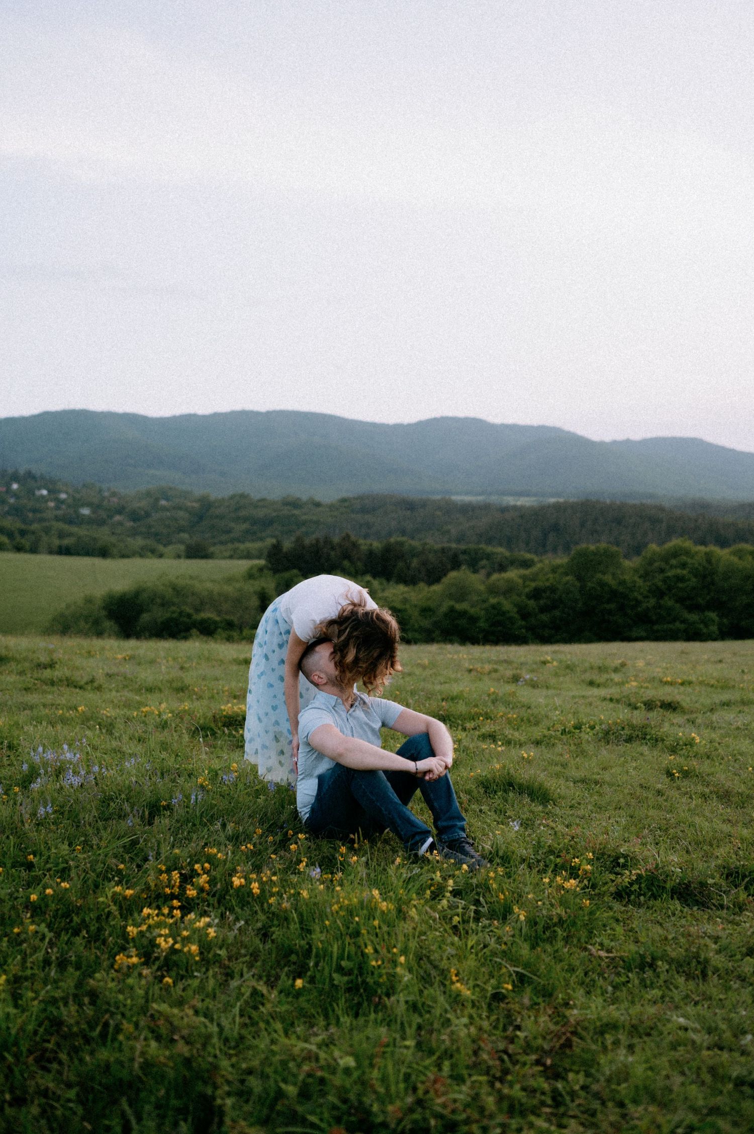 Two people embrace while sitting among wildflowers on a scenic mountain hillside.