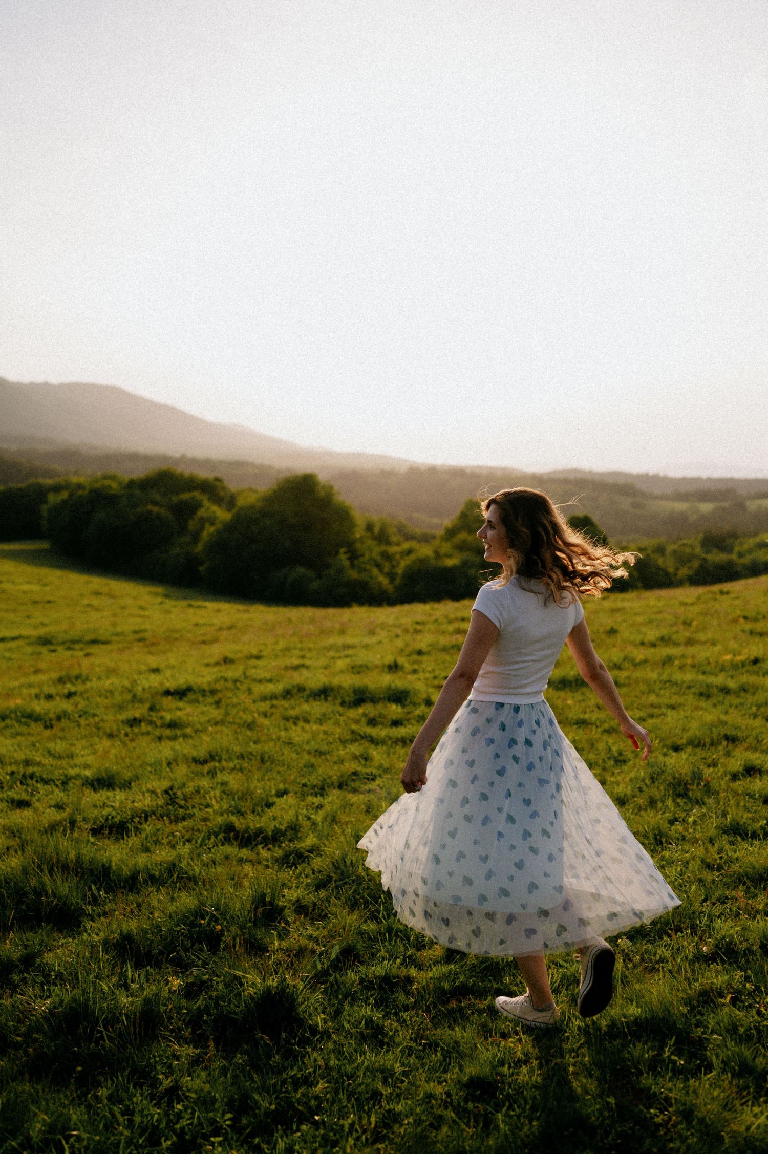 Dancing figure in a white dress spins through tall grass on a scenic hillside.