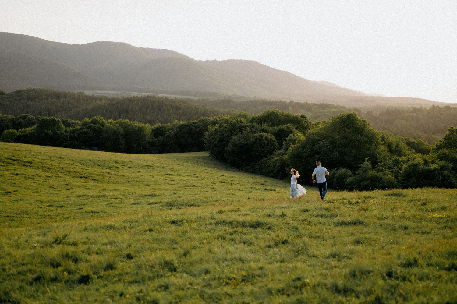 A couple strolls through a lush green field with rolling hills and forest in the distance.
