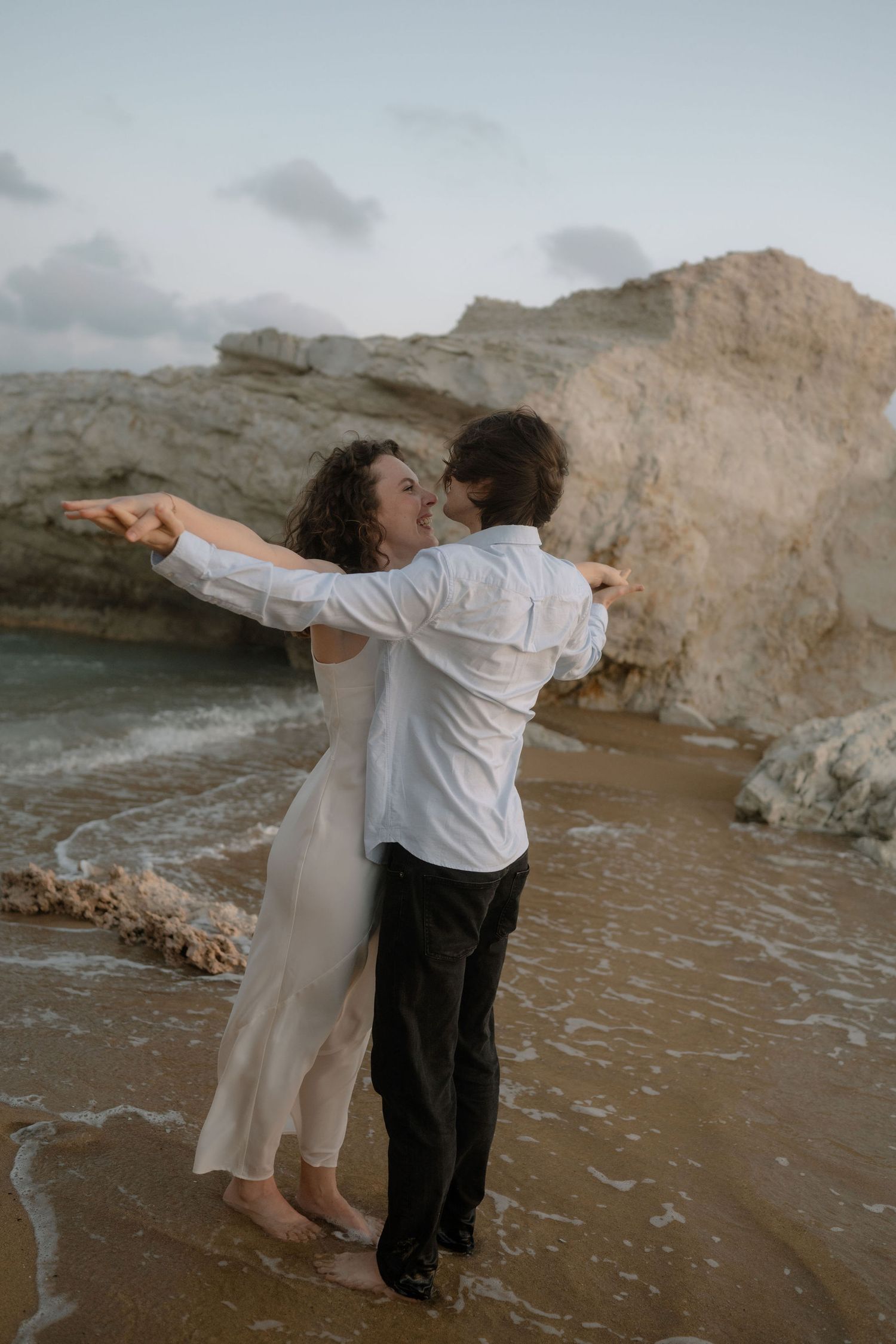 Couple embracing barefoot on a rocky beach shore, woman in white dress with arms outstretched, at dusk.