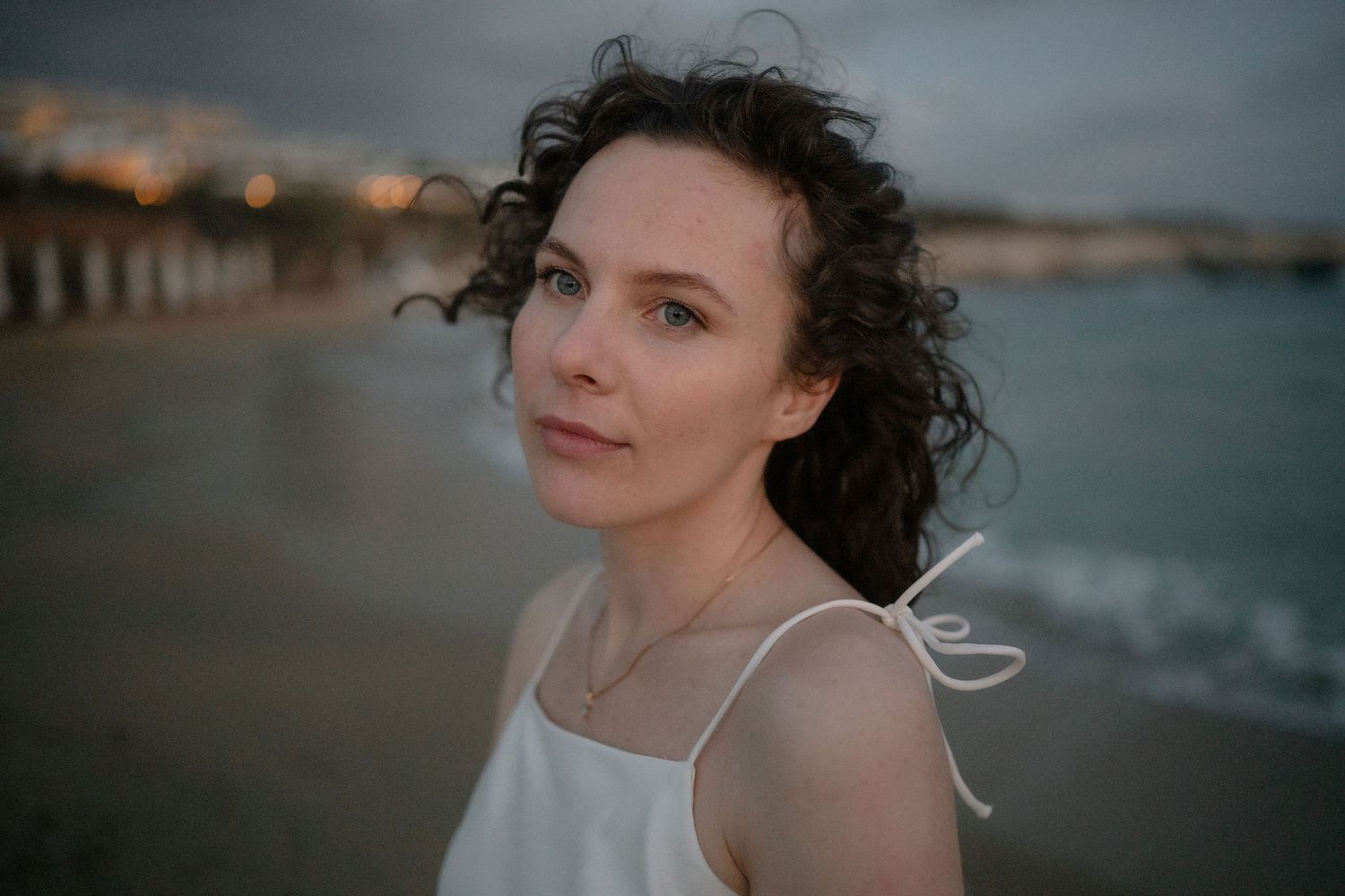 Young woman with curly dark hair in white dress on beach at dusk, ocean waves in background.