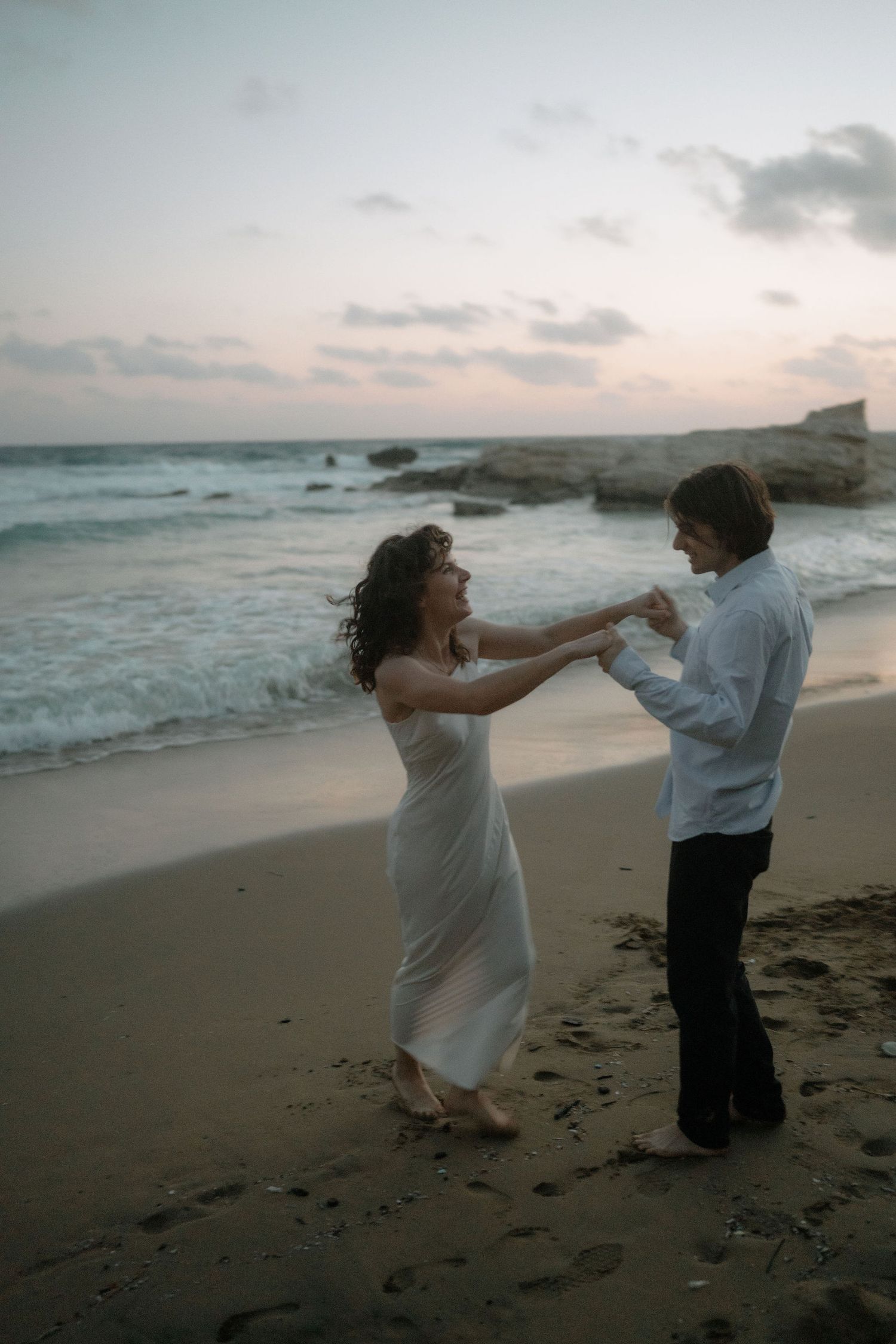 Couple dancing barefoot on sandy beach at sunset, woman in white dress, waves and rocky shore in background.