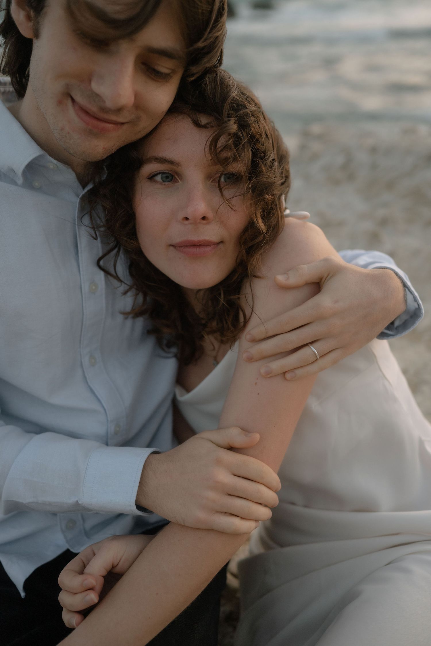A couple shares a tender embrace on a beach, the woman wearing a ring as the man wraps his arms around her.