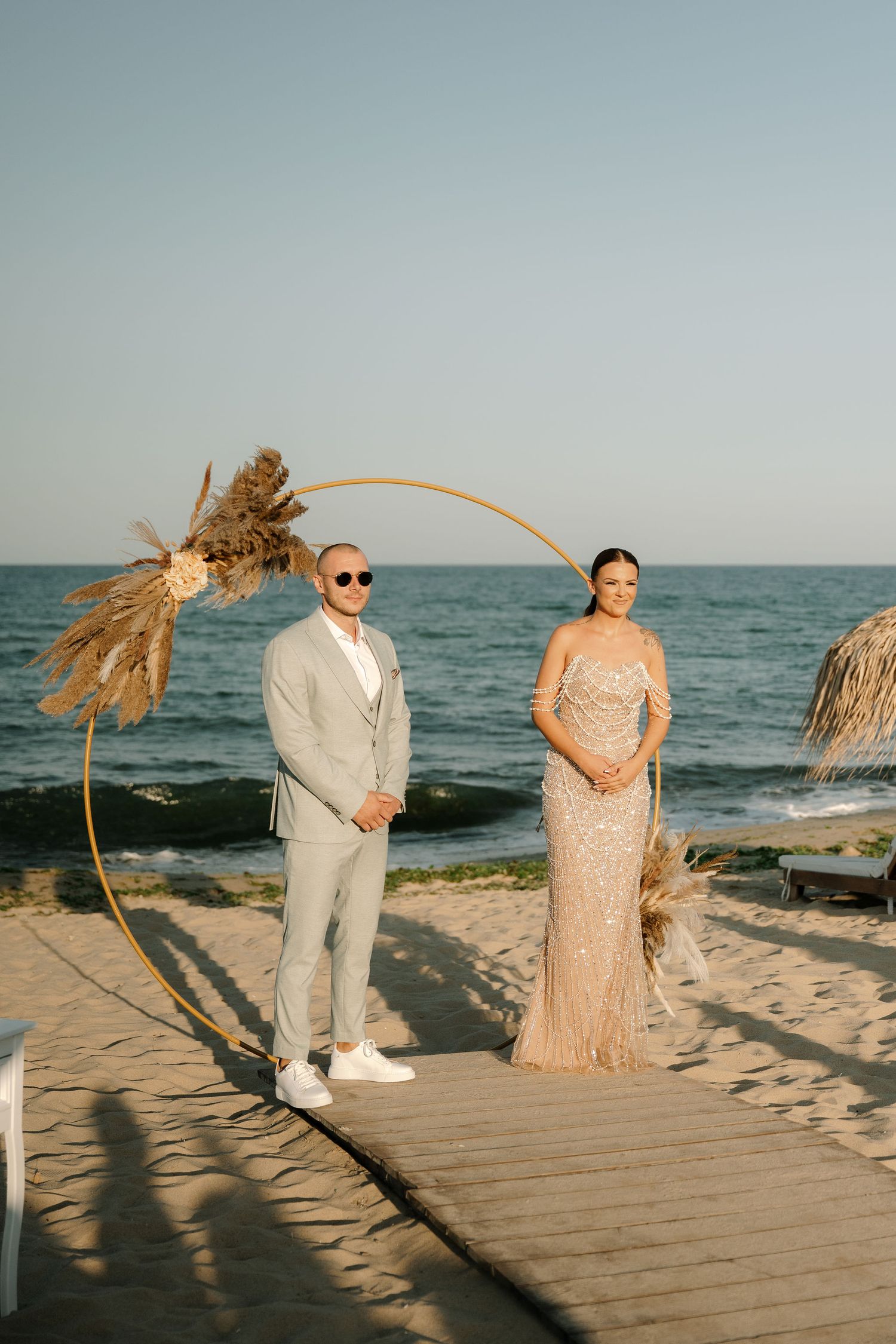 Couple in elegant attire poses together on a wooden boardwalk at sunset by the beach.