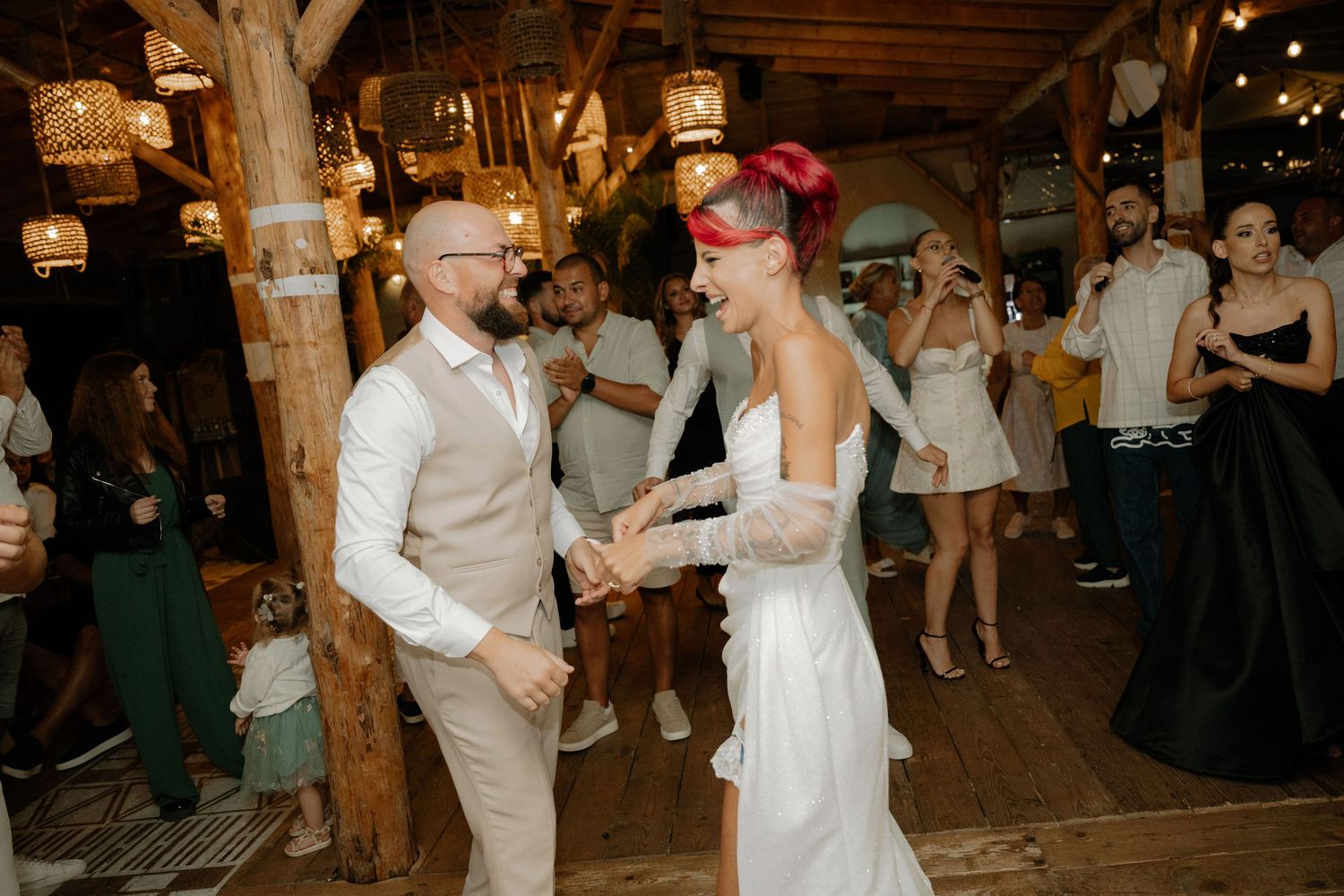 A couple in wedding attire dances together on a wooden floor surrounded by guests at an evening reception.