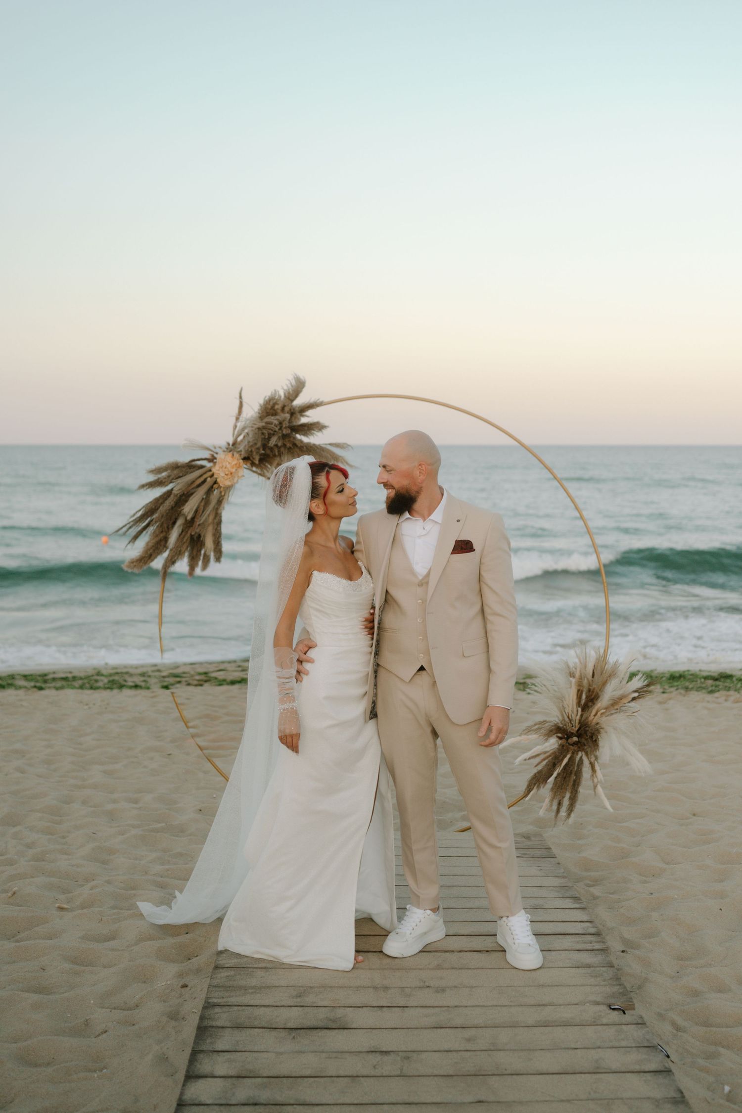 A couple poses for romantic beach wedding photos at sunset with a palm-decorated arch on a wooden boardwalk.
