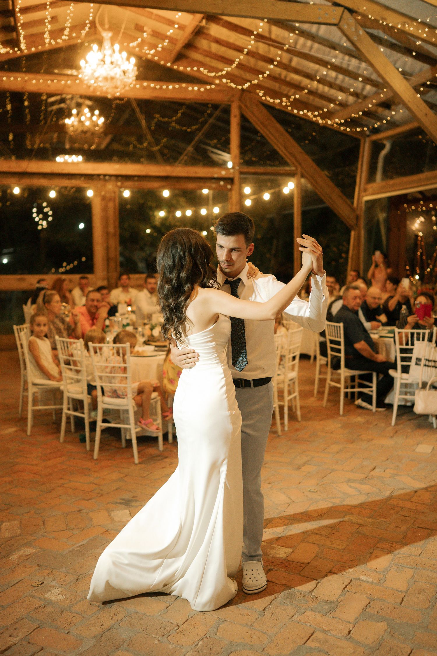 Couple performs romantic dance moves during their first dance at rustic barn wedding reception with string lights overhead.