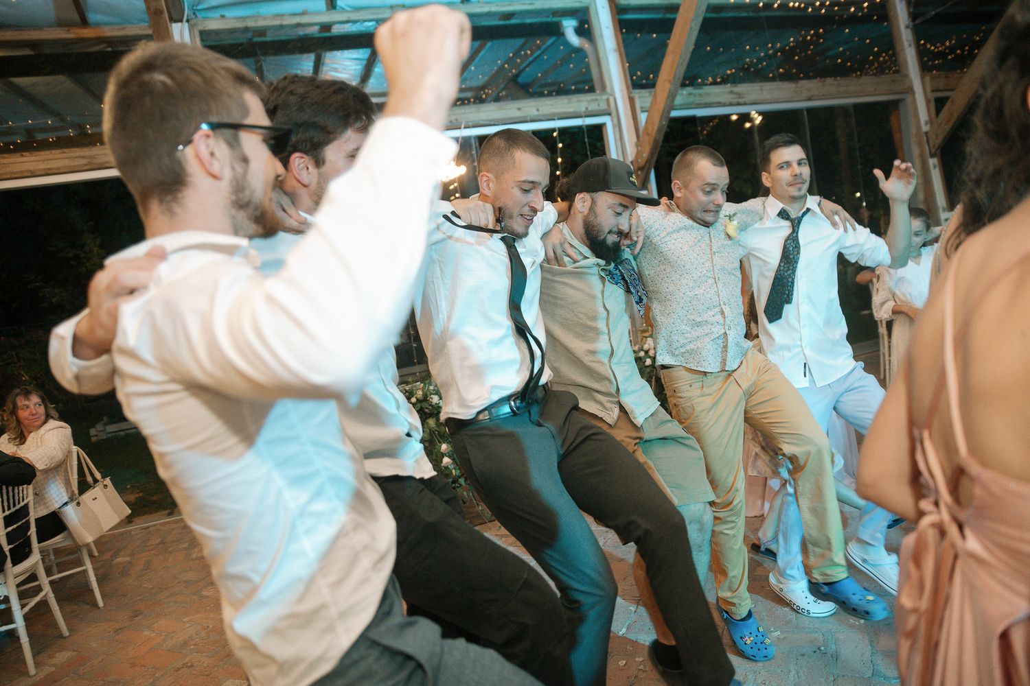 Groomsmen in suspenders and dress shirts dancing in a line at a rustic wedding reception venue.
