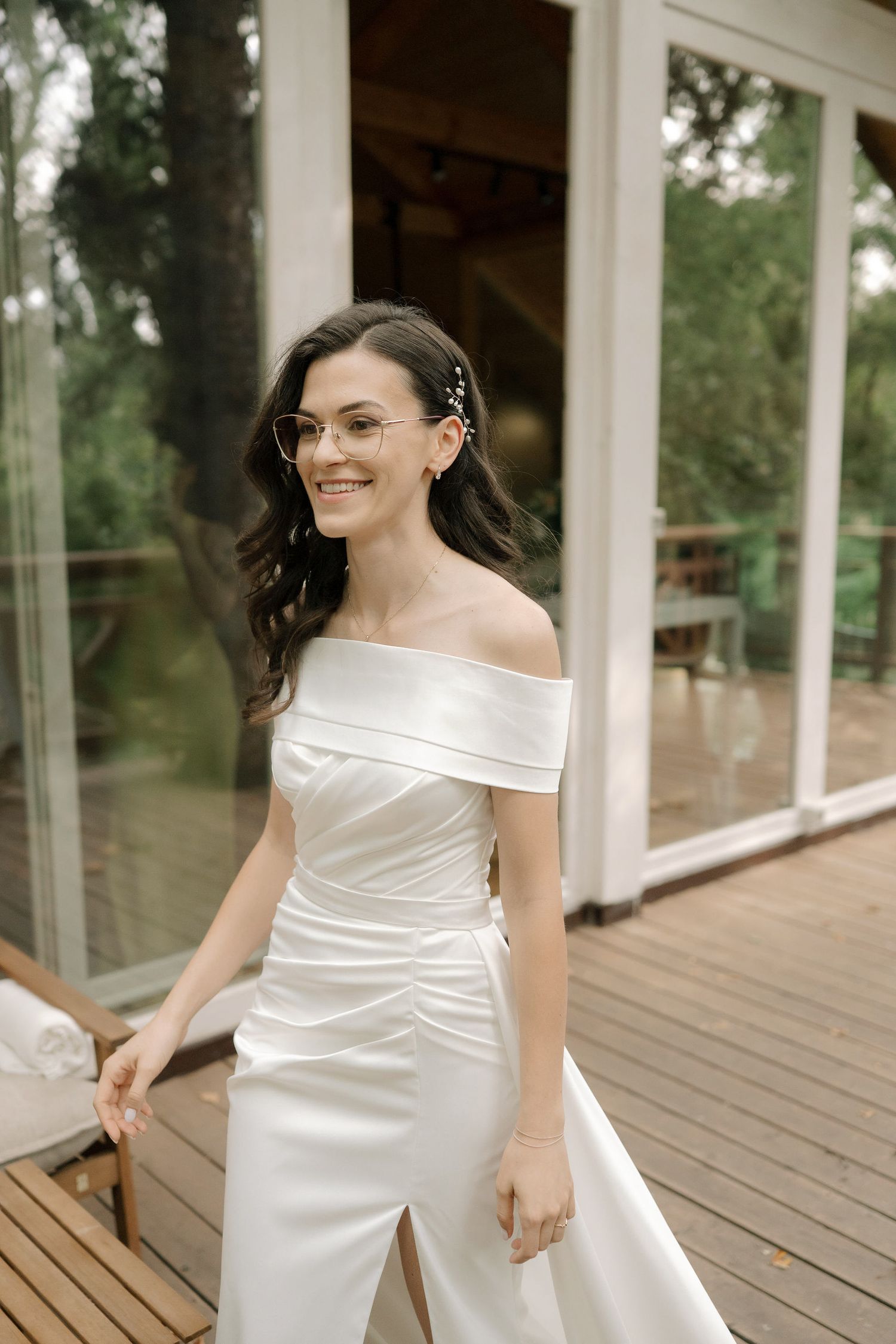 A bride in an elegant off-shoulder white gown poses gracefully on a wooden deck surrounded by glass windows.