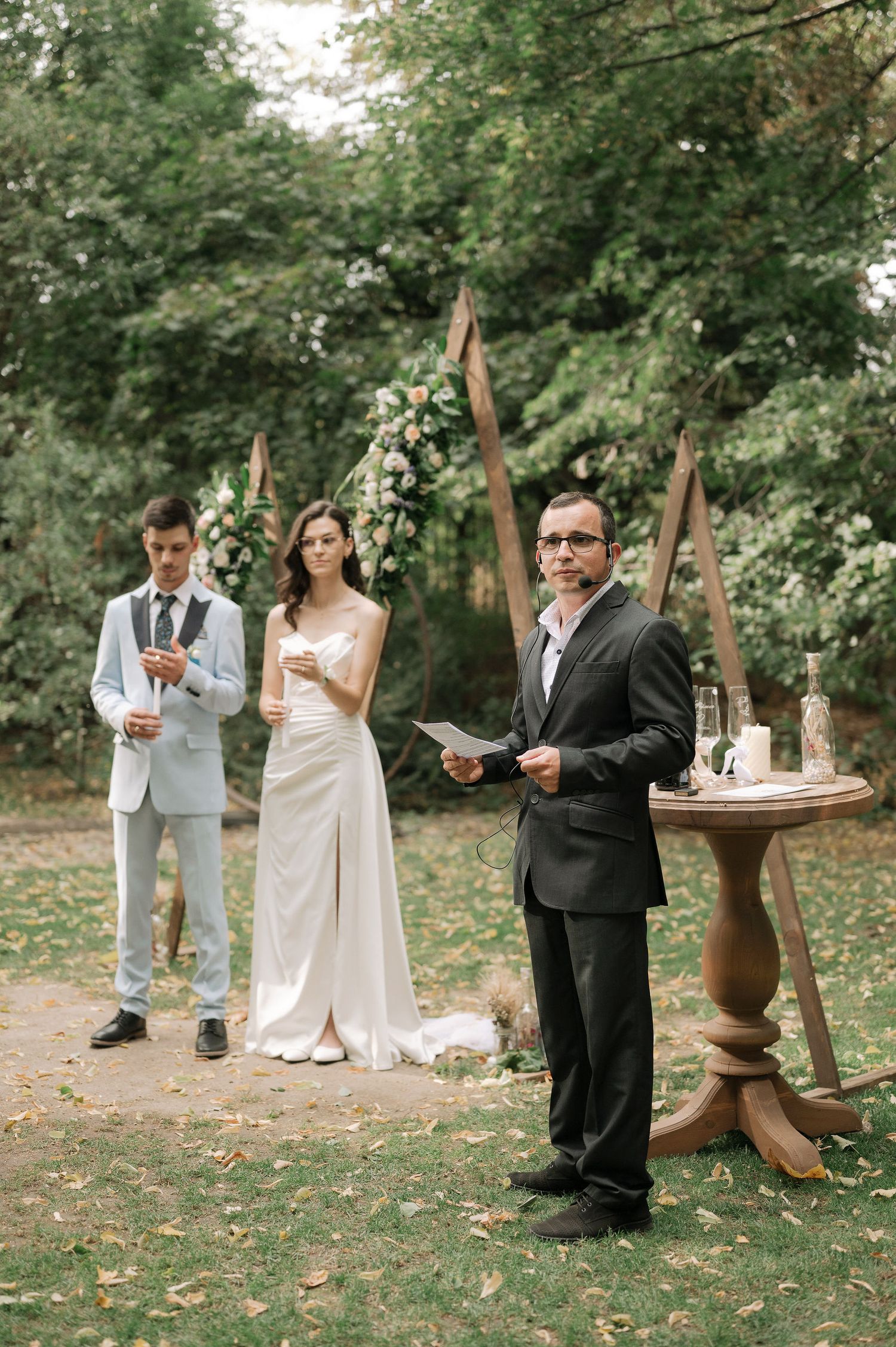 A wedding party gathers for a toast in a serene outdoor garden setting with scattered flower petals on the ground.
