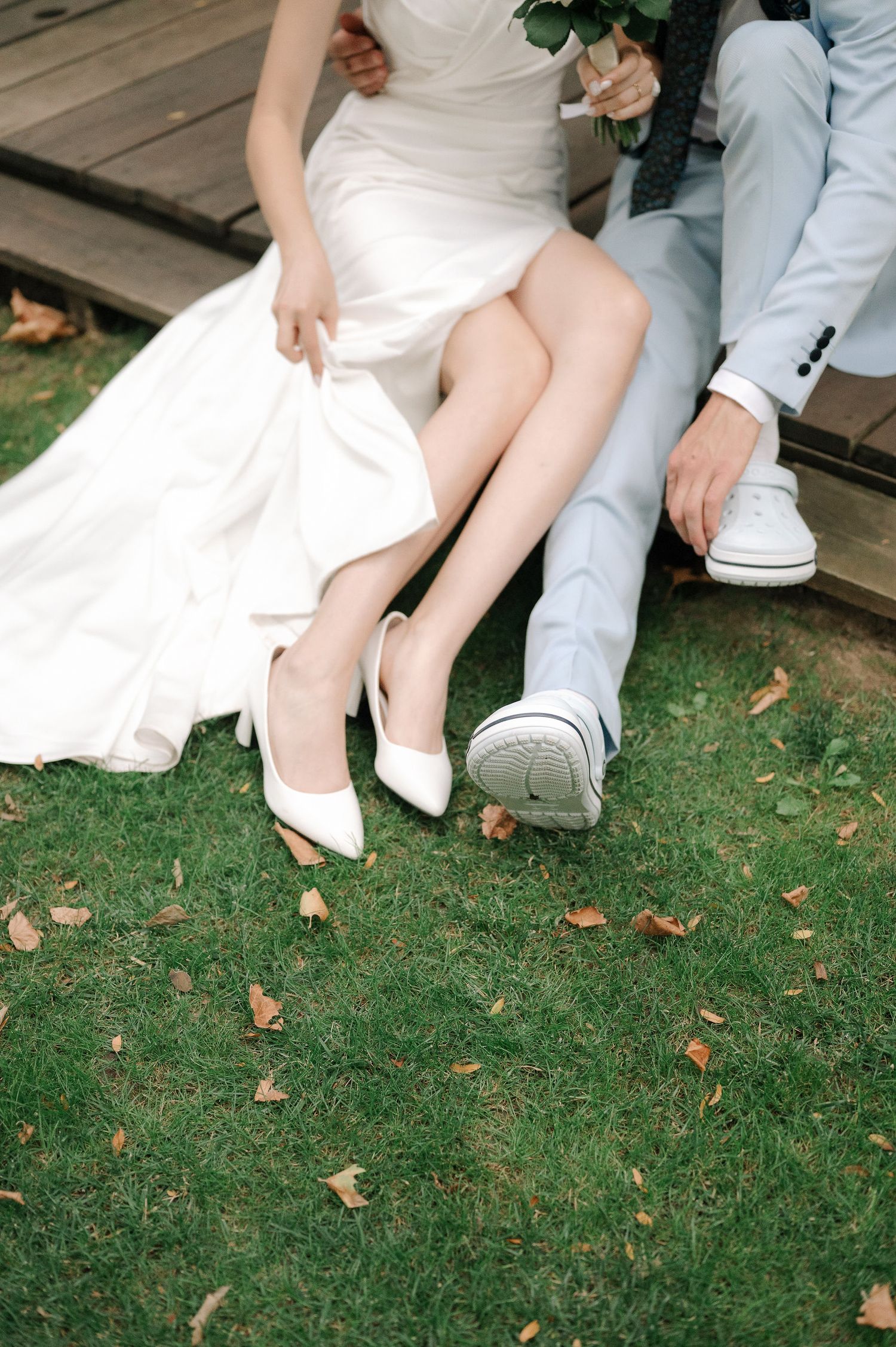 Bride in white dress and groom in light suit sit on wooden deck showing their matching white shoes on grass with fallen leaves.