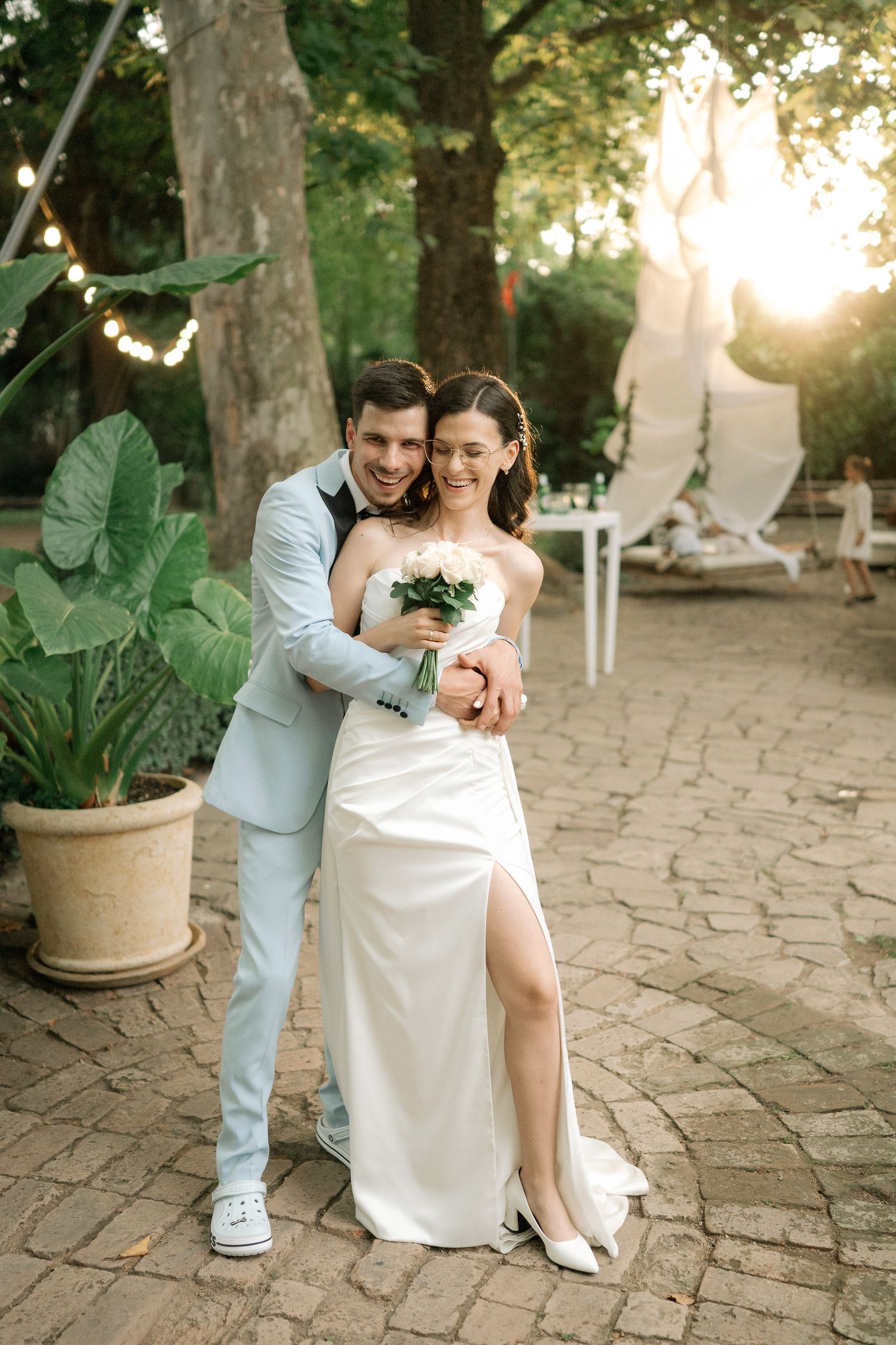 A romantic couple in wedding attire sits together on stone pavers in a garden setting during golden hour.