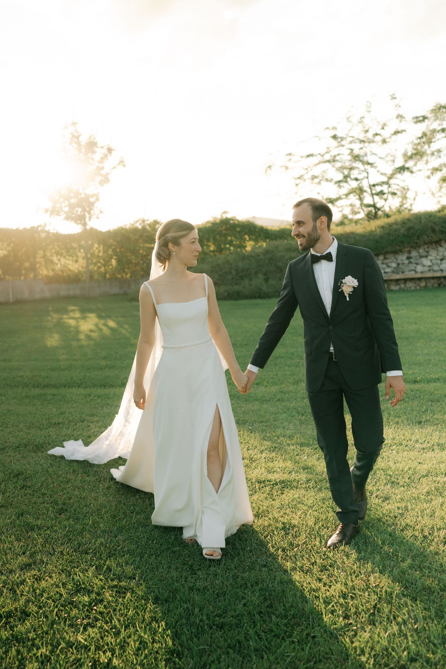 Newlywed couple walks hand in hand across a sunlit grassy field during golden hour at their outdoor wedding.