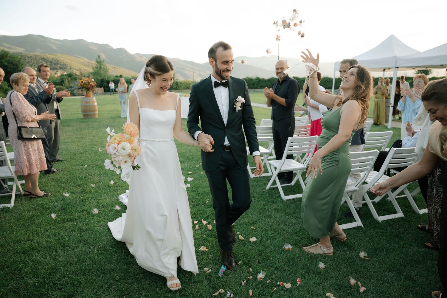 A joyful wedding celebration on a grassy lawn with mountains in the background as guests throw flower petals.
