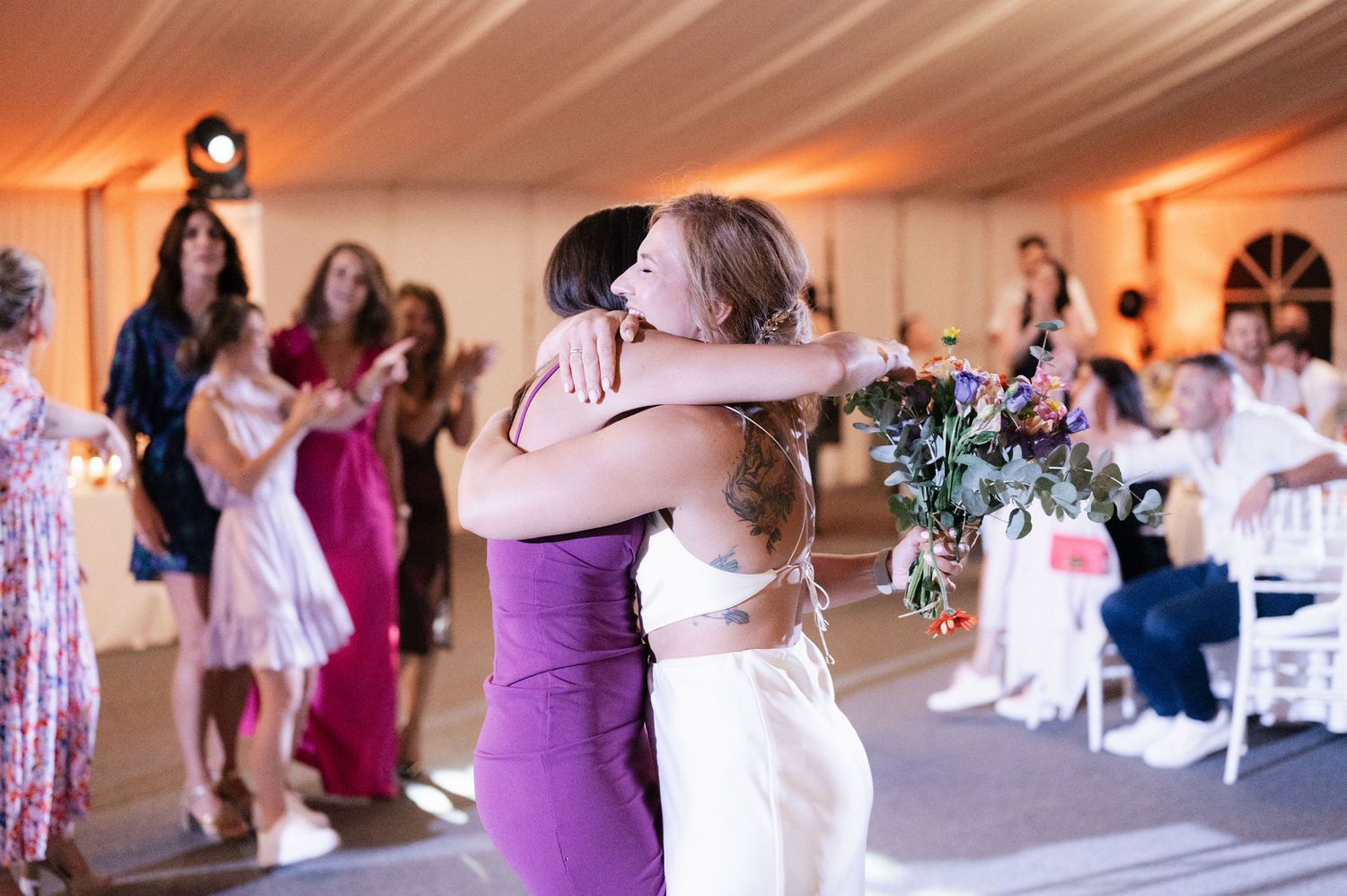 Wedding guests dance and celebrate on a dance floor under warm lighting at an indoor reception venue.