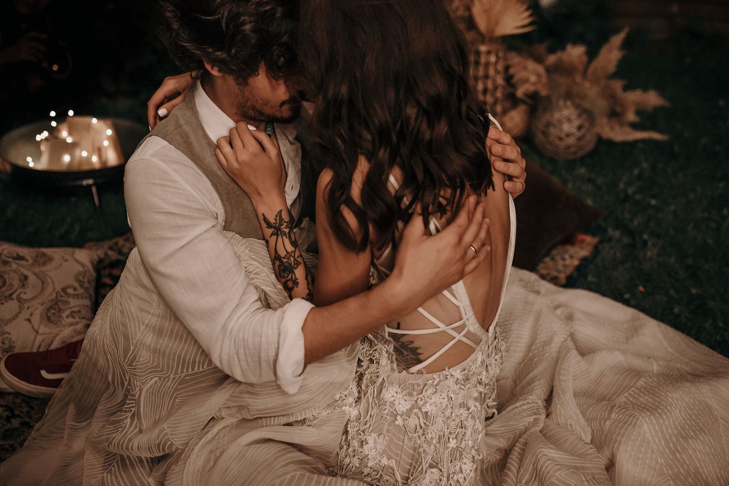 Intimate couple sitting on grass at dusk with twinkling lights in background wearing formal attire.