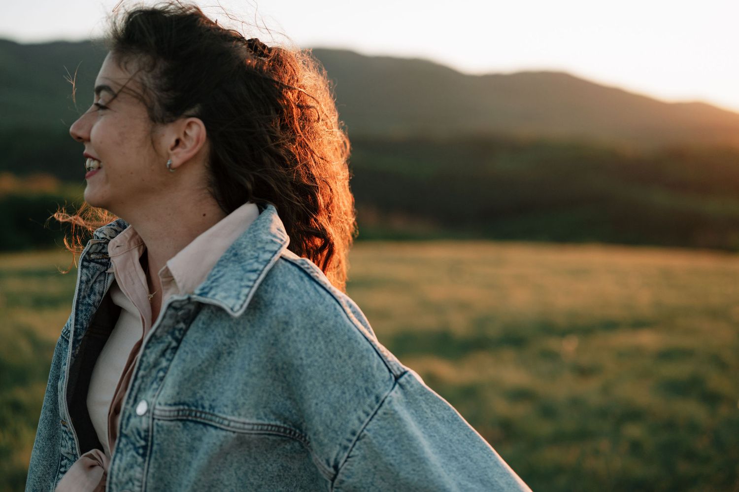 Person with curly hair in denim jacket looking at sunset over golden field during magic hour.