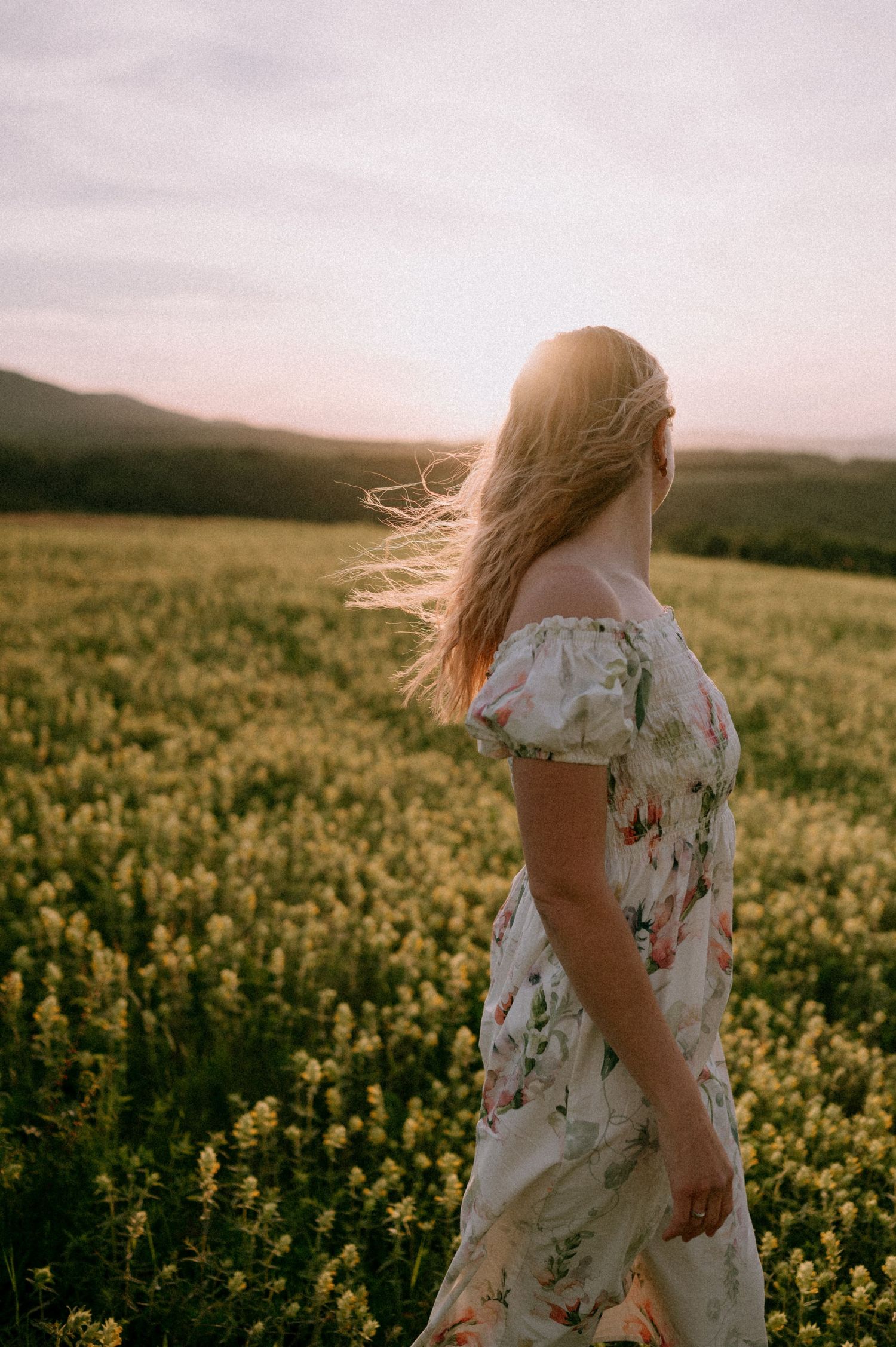 Person in white floral dress standing among blooming yellow wildflowers at dusk.