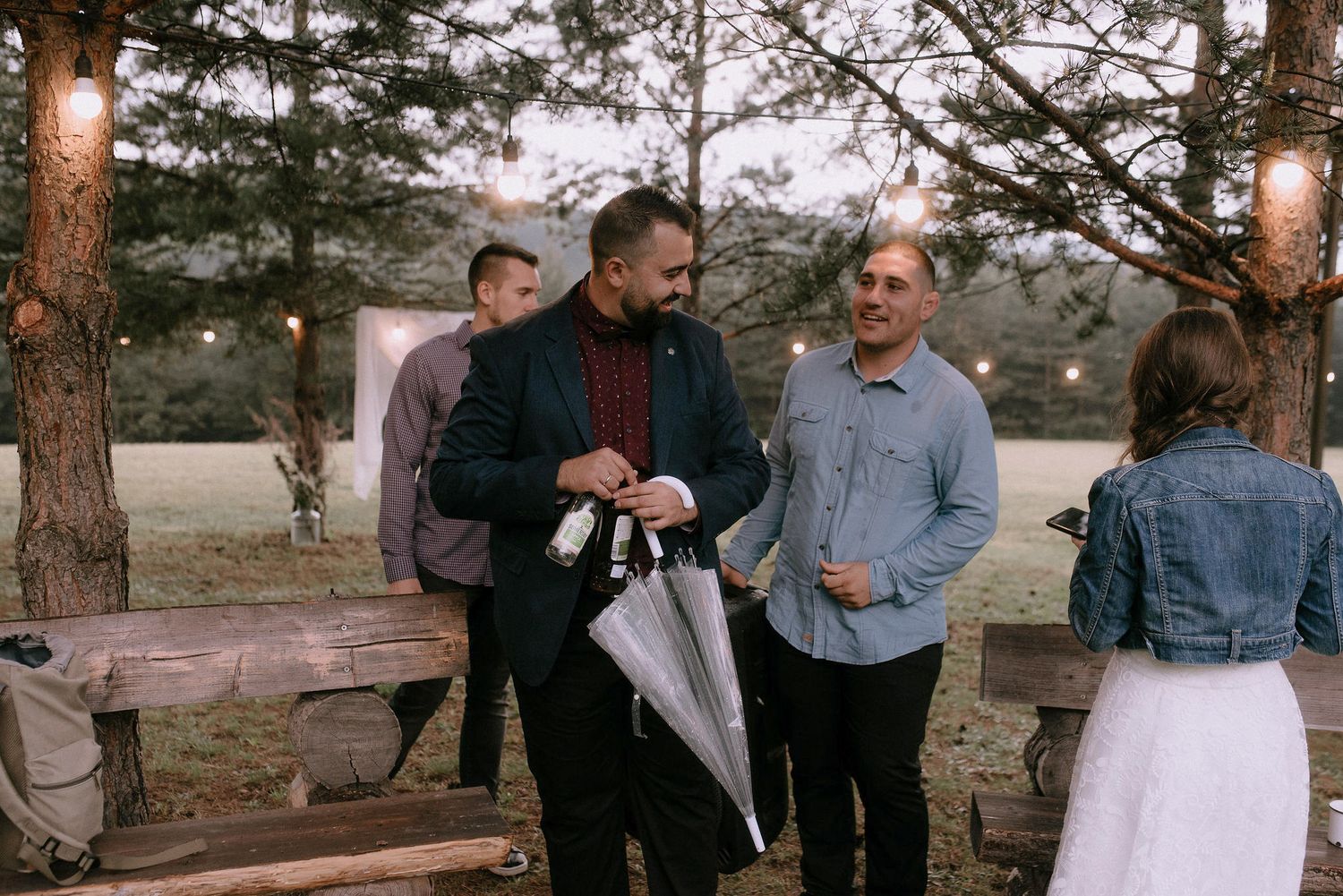 Wedding guests gather outdoors in a pine forest setting during golden hour.