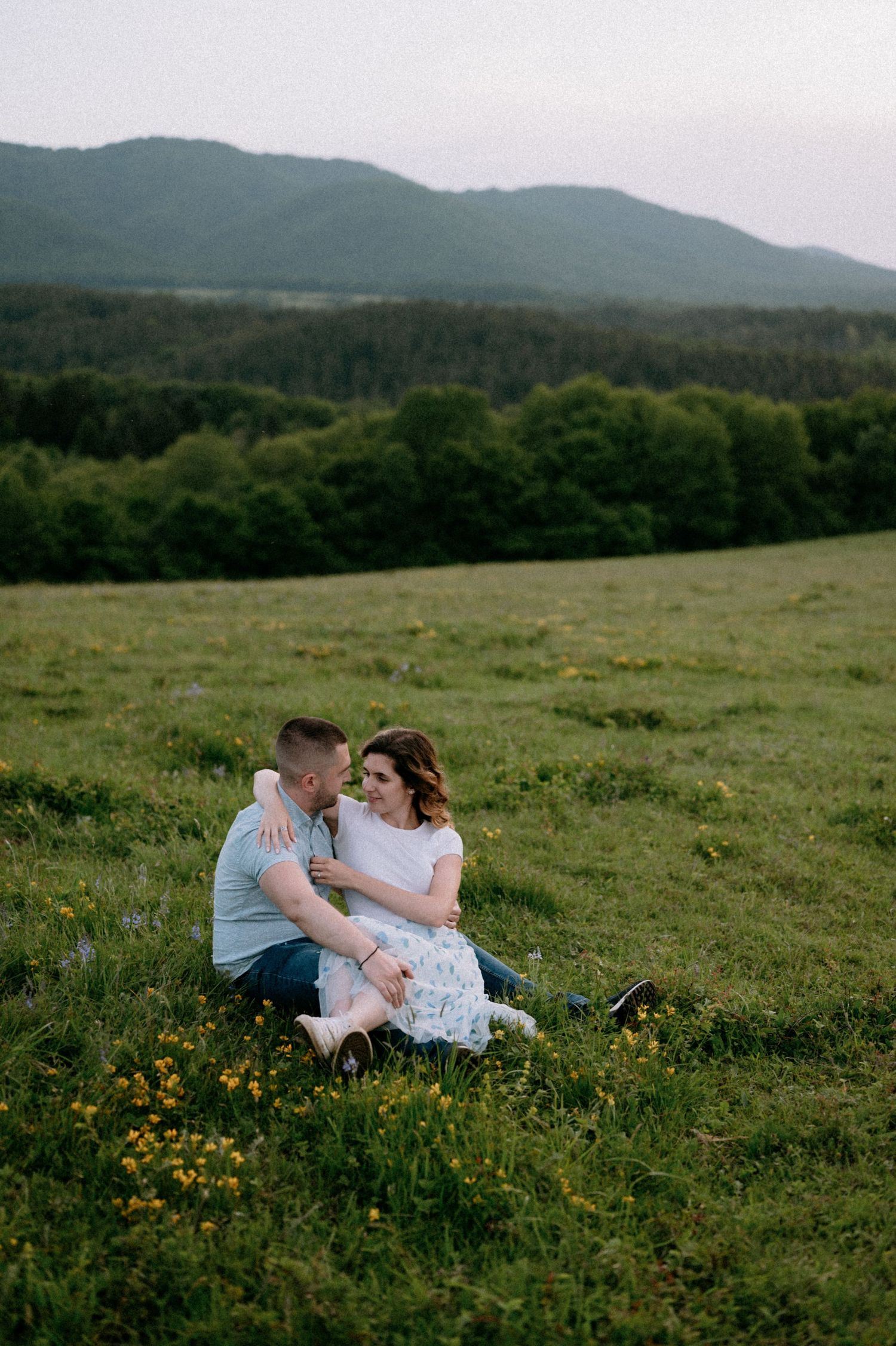 A romantic couple sits together in a wildflower meadow with rolling mountains in the background at sunset.
