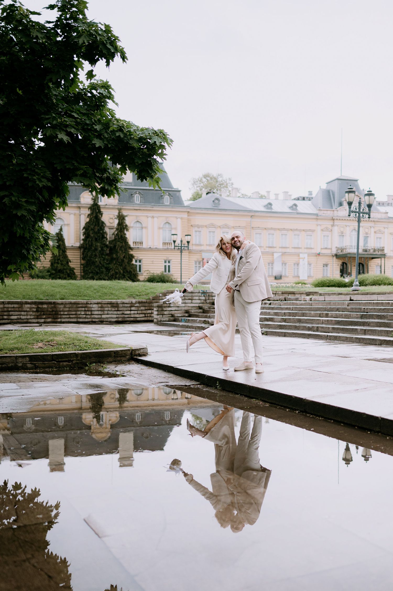 Elegant couple performing dance moves by large reflecting pool at historic European palace grounds.