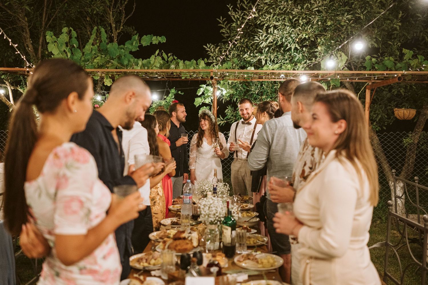 Outdoor evening wedding reception with string lights illuminating guests mingling at tables.