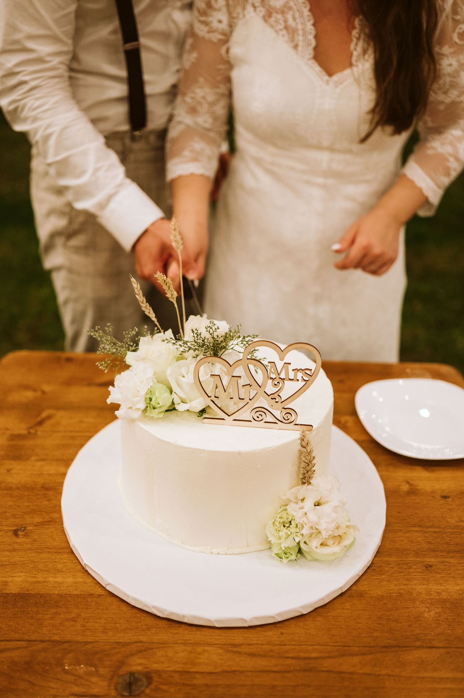Couple cutting their elegant white wedding cake decorated with flowers and a wooden topper.
