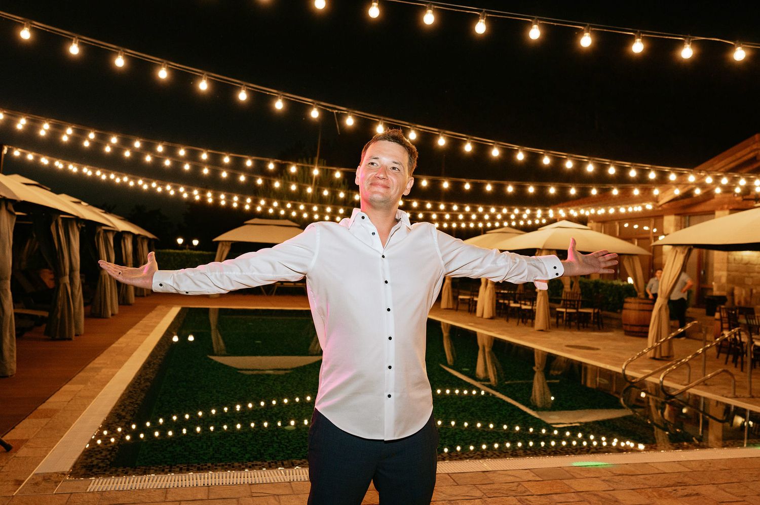 Person poses with outstretched arms under string lights at an outdoor evening event.