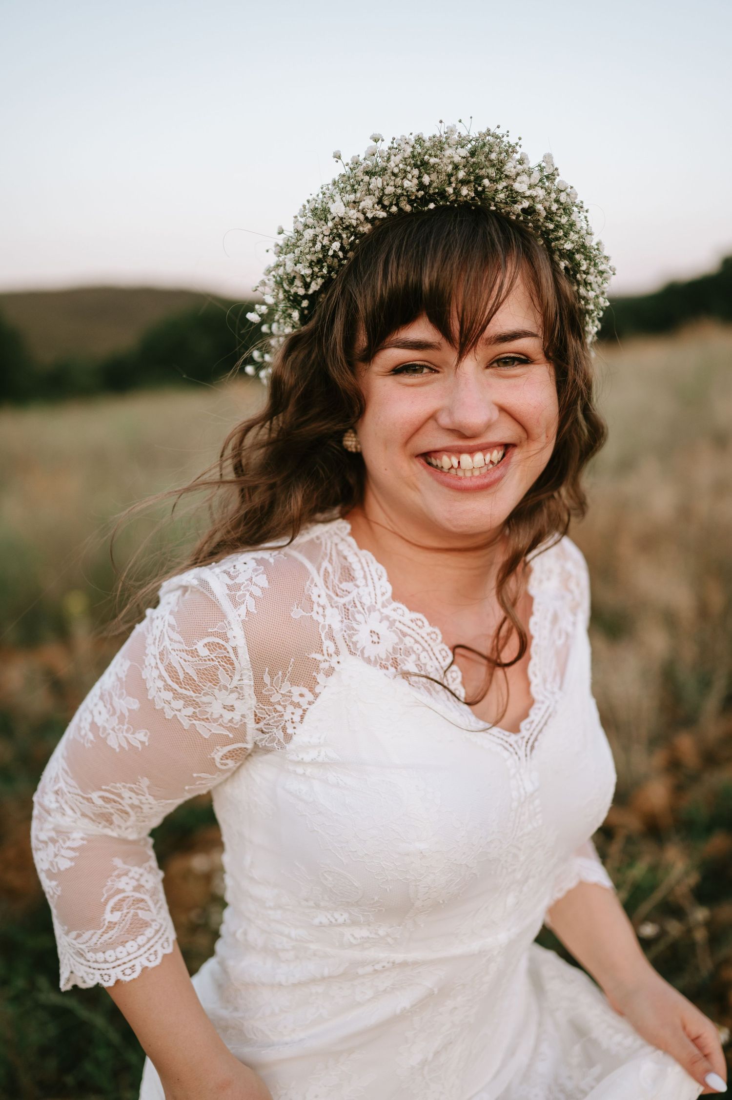 Bride wearing delicate lace dress and floral crown in natural outdoor setting.