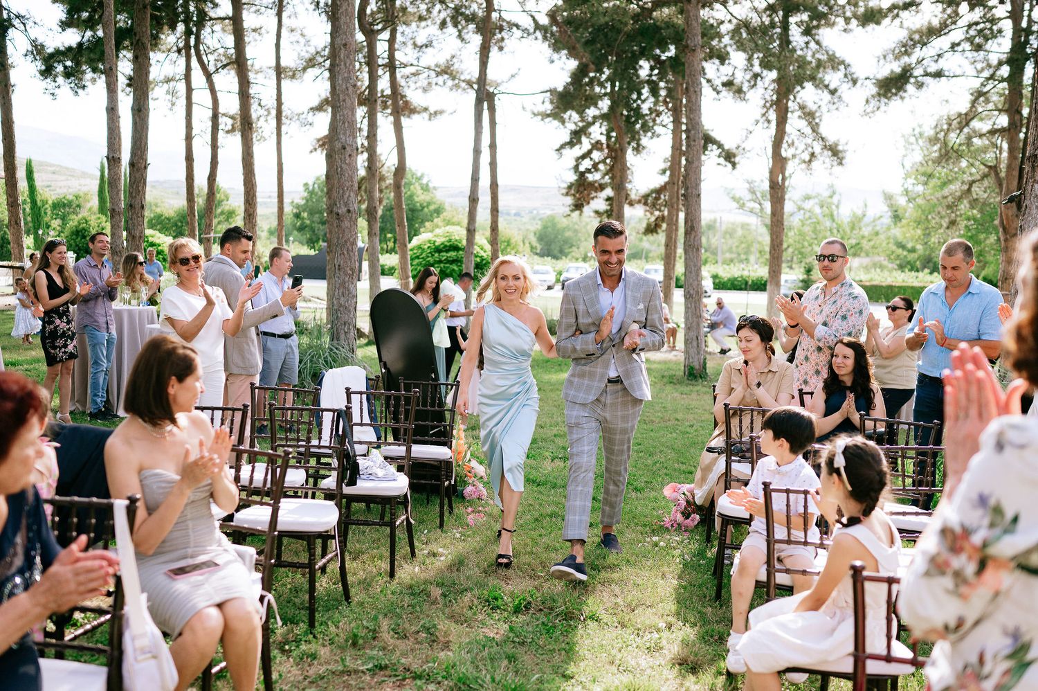 Wedding guests applauding during outdoor ceremony processional among tall pine trees.