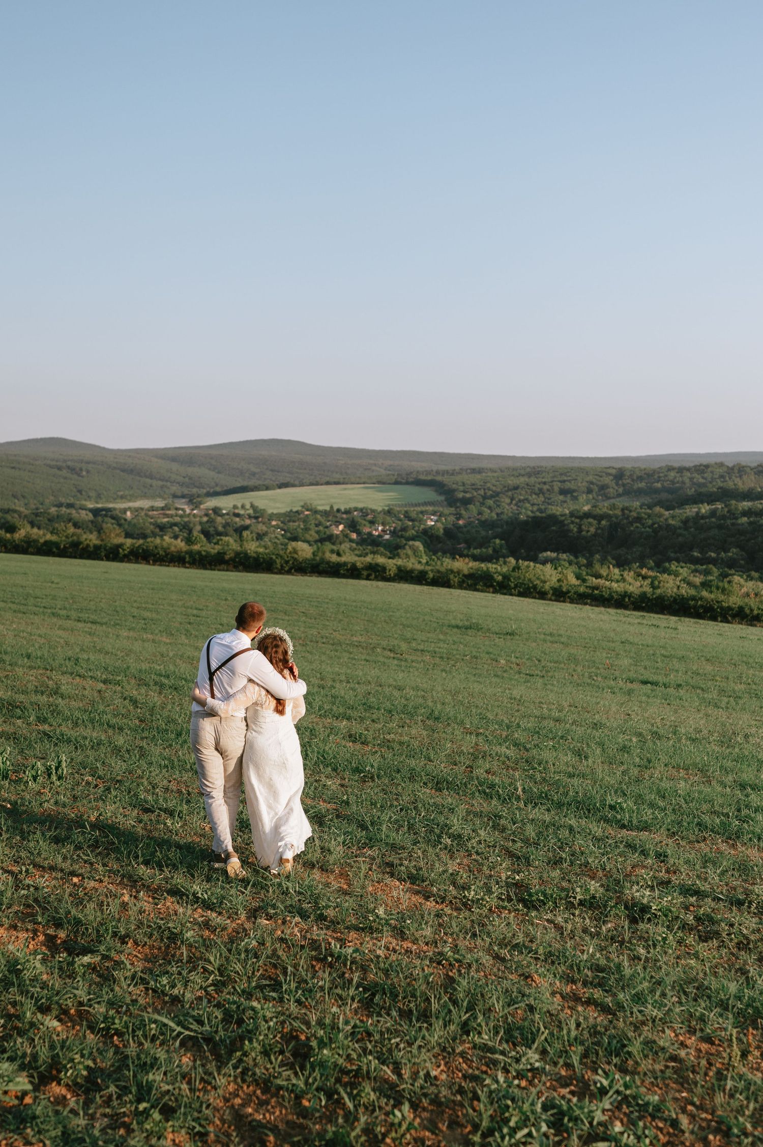 A couple in white attire walks together across a scenic hilltop meadow.