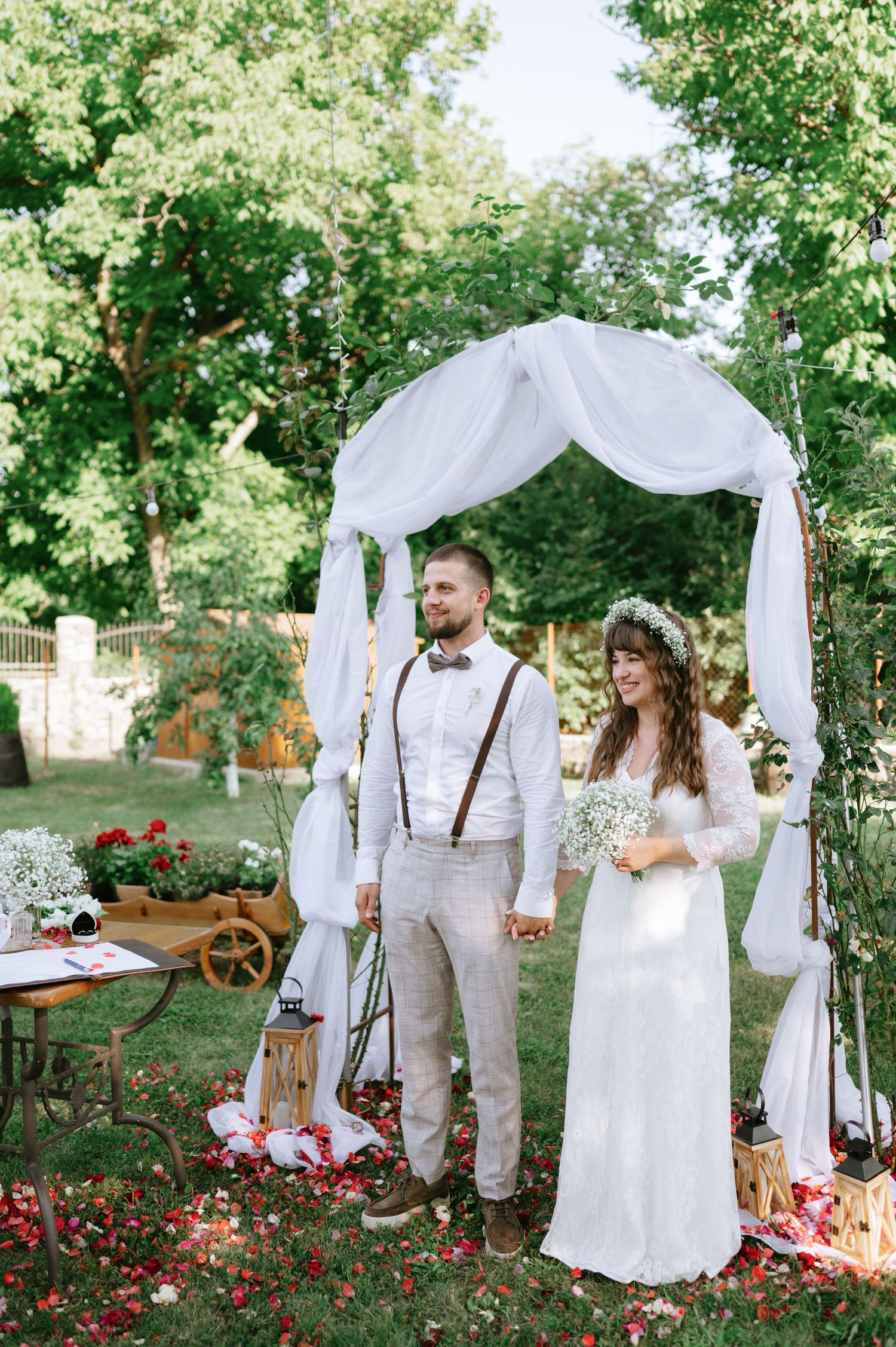 A romantic outdoor wedding ceremony setup with white drapes and red flowers in a garden setting.